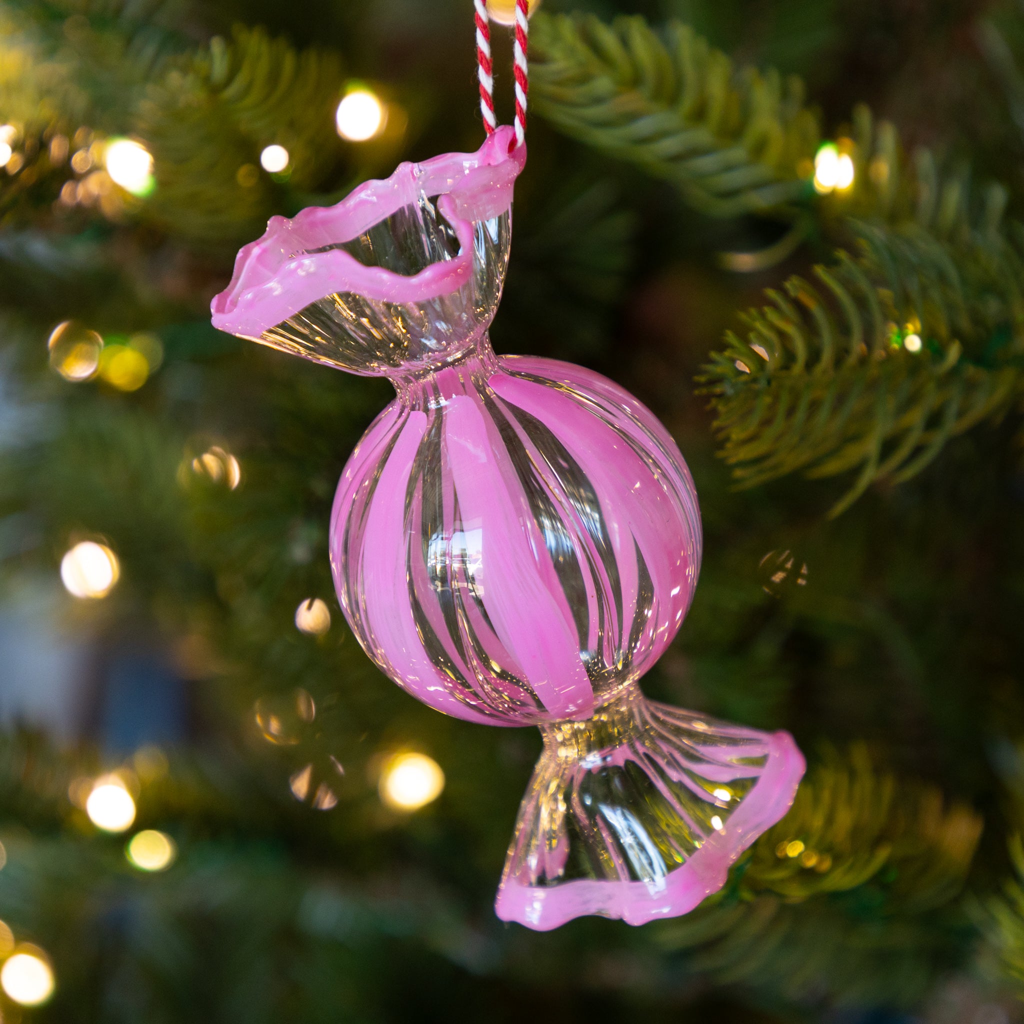 Round, pink glass candy ornament hanging on a Christmas tree with blurred lights in the background.