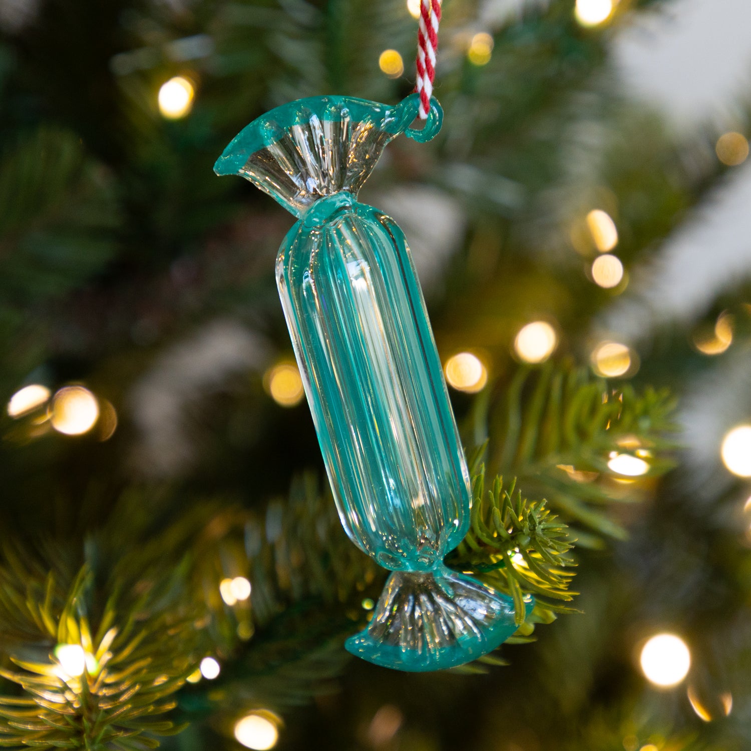 Long teal candy-shaped ornament on a Christmas tree with blurred lights in the background.