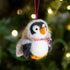 Felted penguin ornament with a red and white striped scarf against a blurred Christmas tree background.