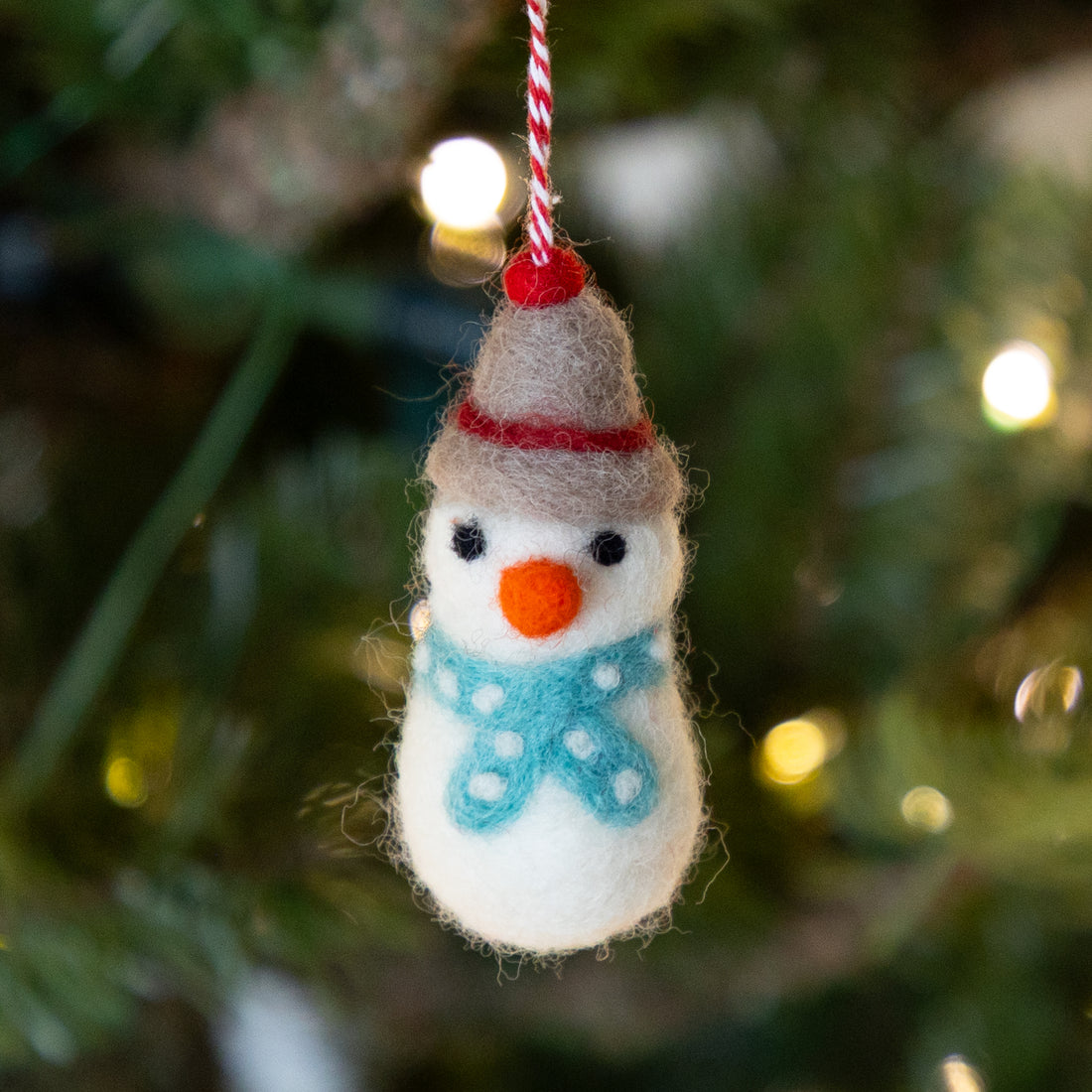 Felt snowman ornament hanging on a Christmas tree with blurred lights in the background.