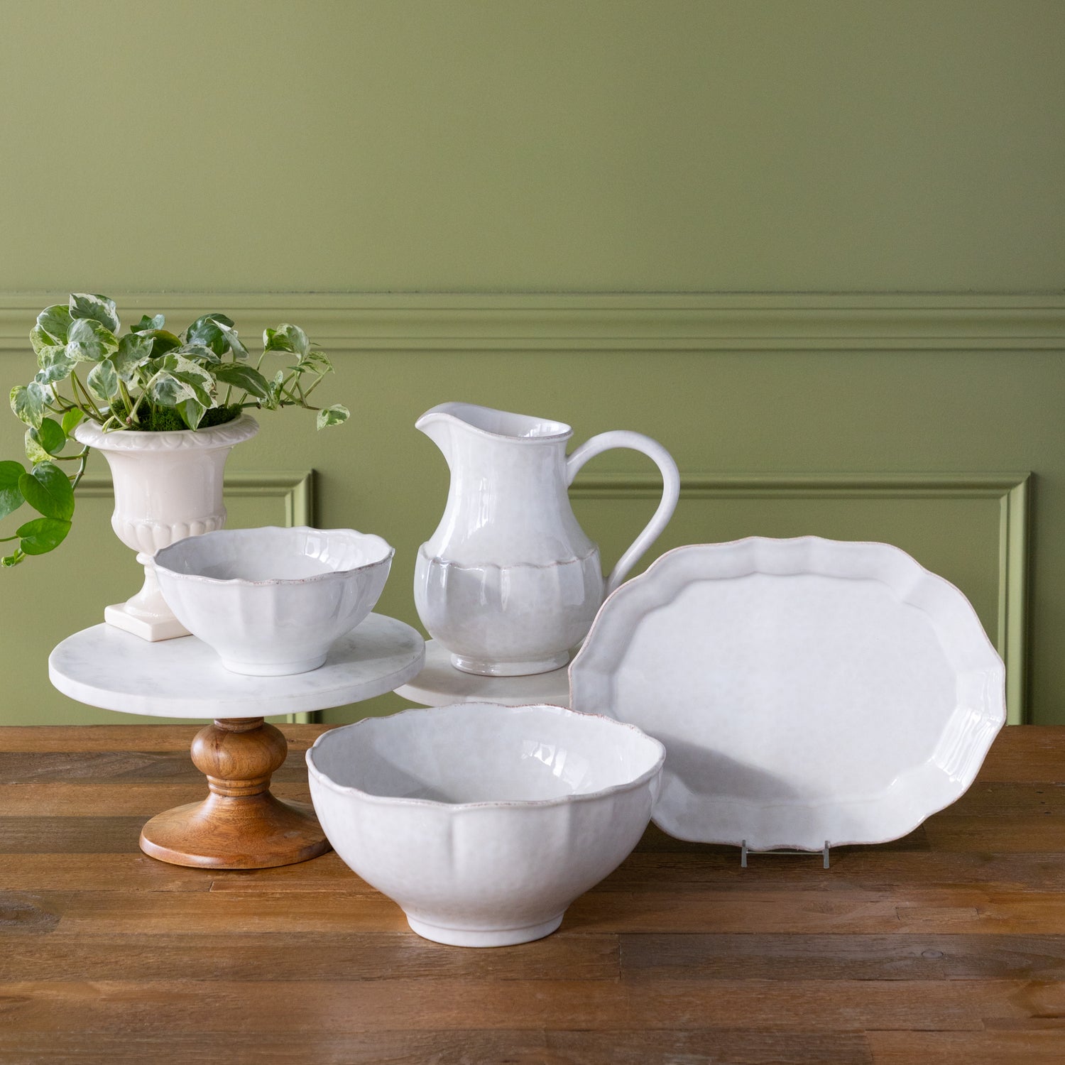Set of white ceramic serveware including bowls, a pitcher and platter on a wooden table with a green wall background.