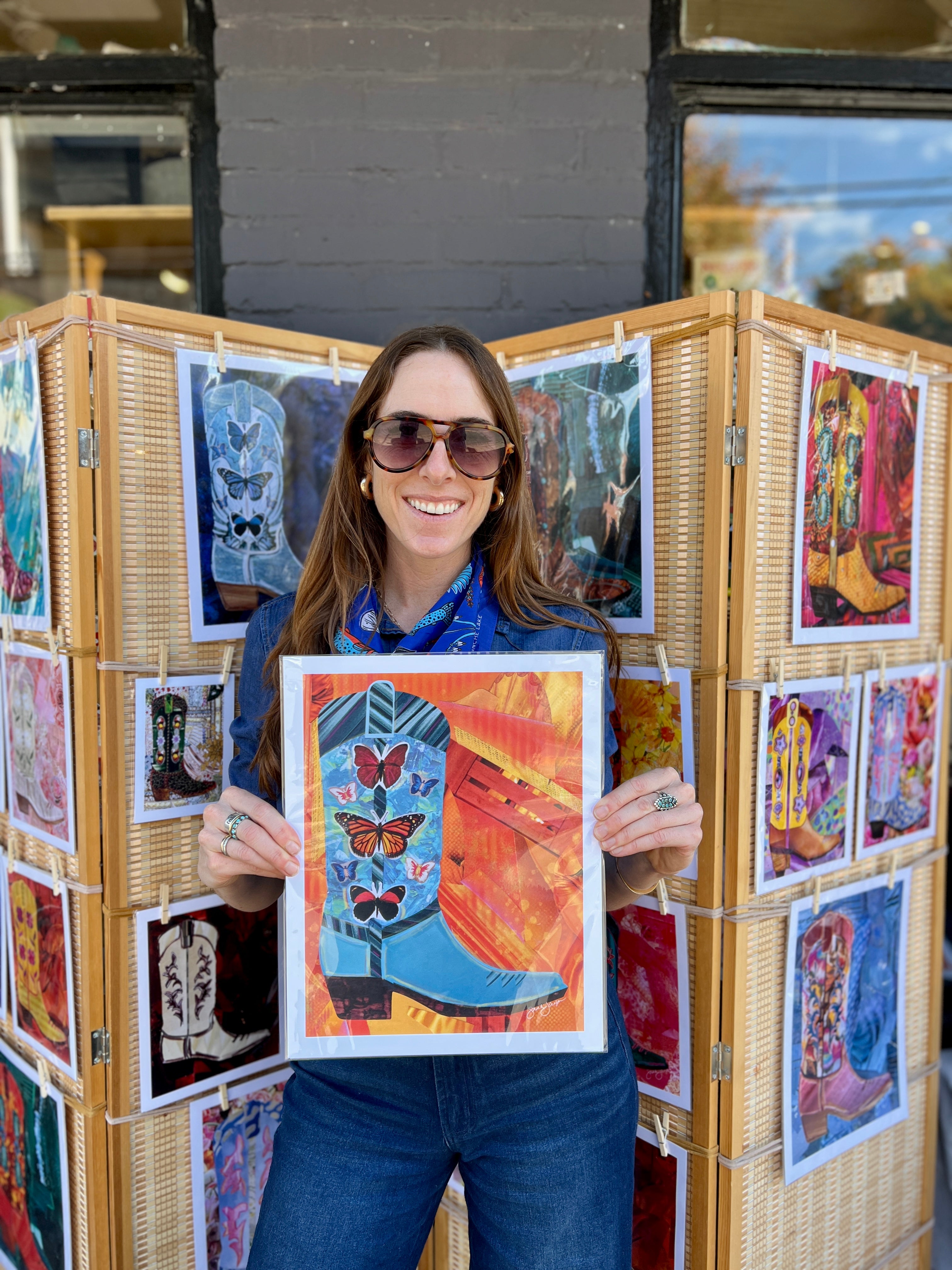 Woman holding a framed artwork of a boot with butterflies, standing in front of a display of other artworks.