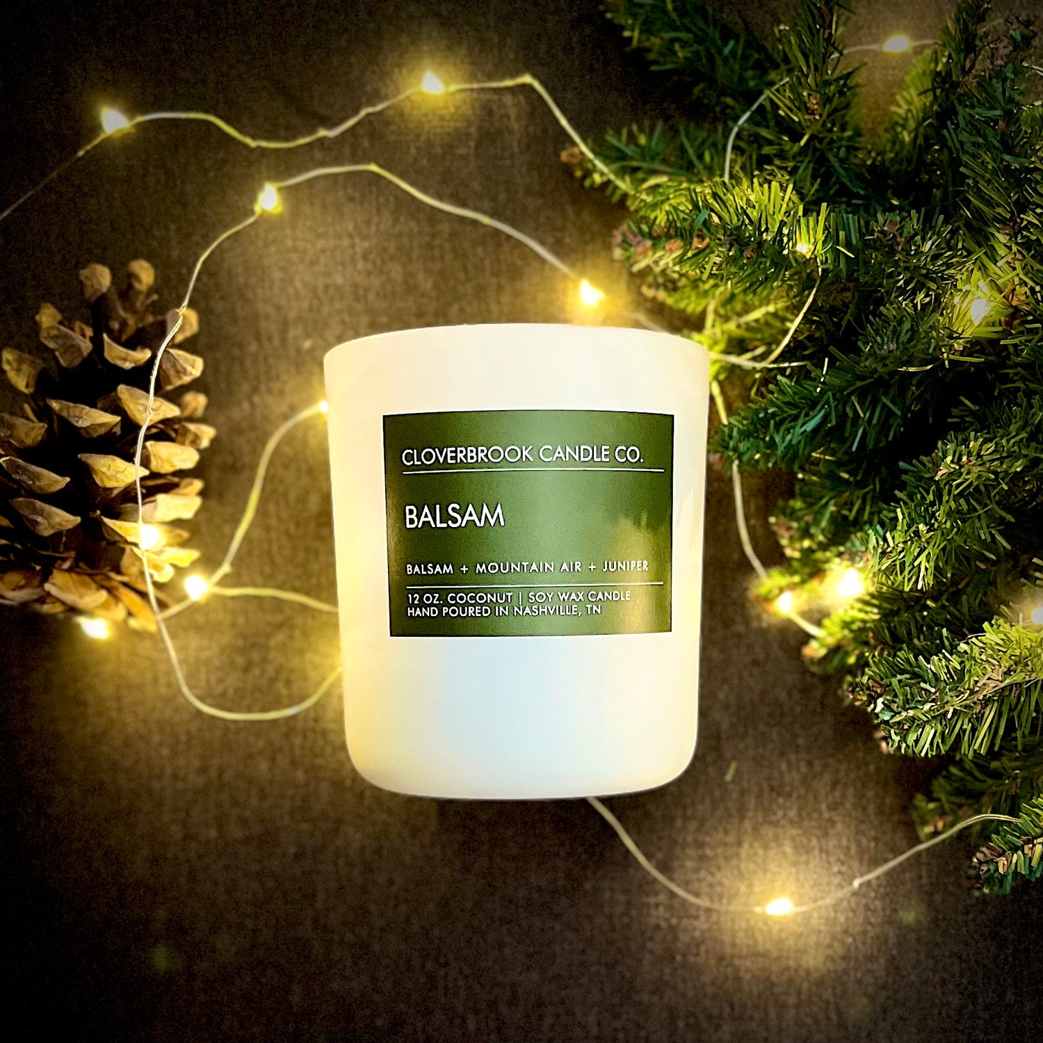 Cloverbrook Candle Co., Balsam candle styled with a pinecone, a Christmas tree branch and lights.