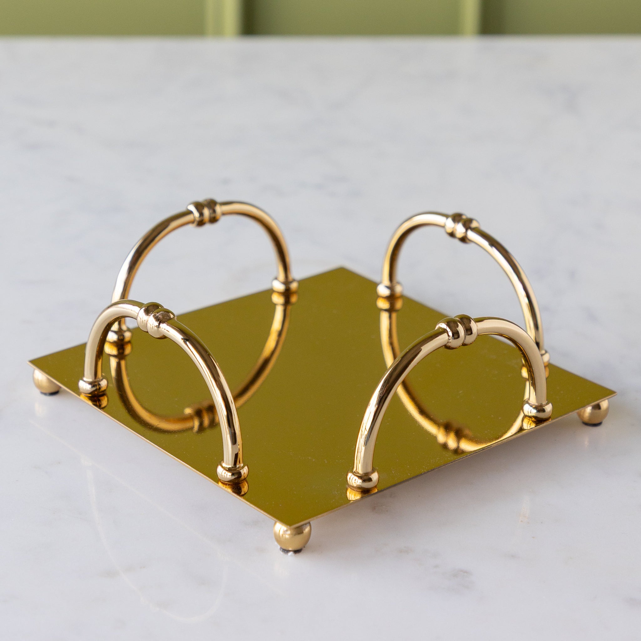 A gold square metal napkin holder with four arched handles sits on a white marble surface.