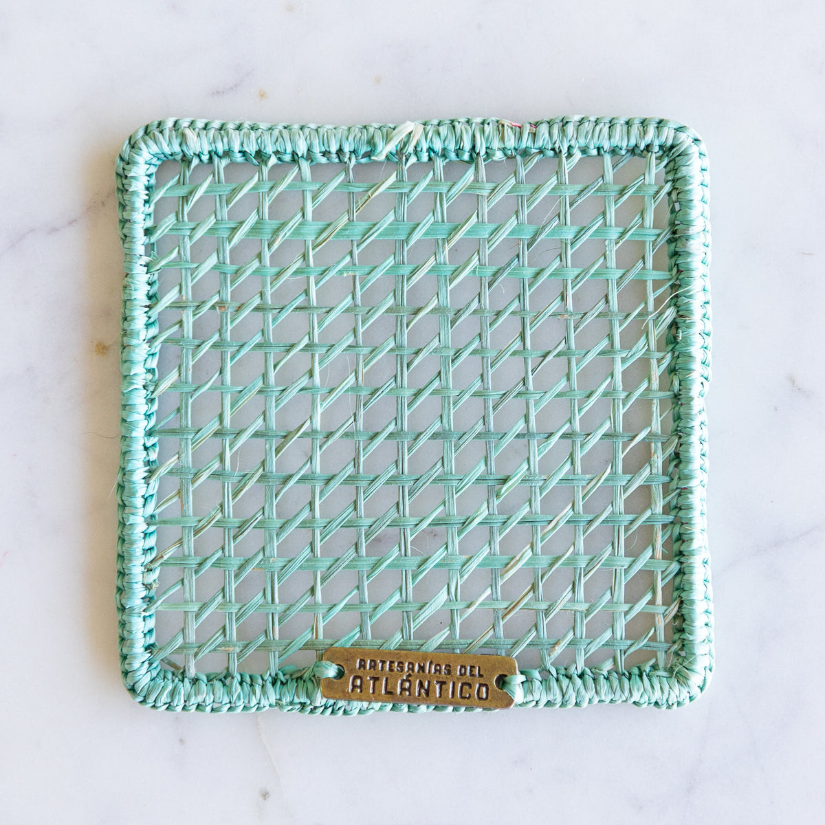 A square woven rattan coaster with a mint green border and a small brass metal tag at the bottom . The coaster is photographed flat-lay style on a white marble surface.