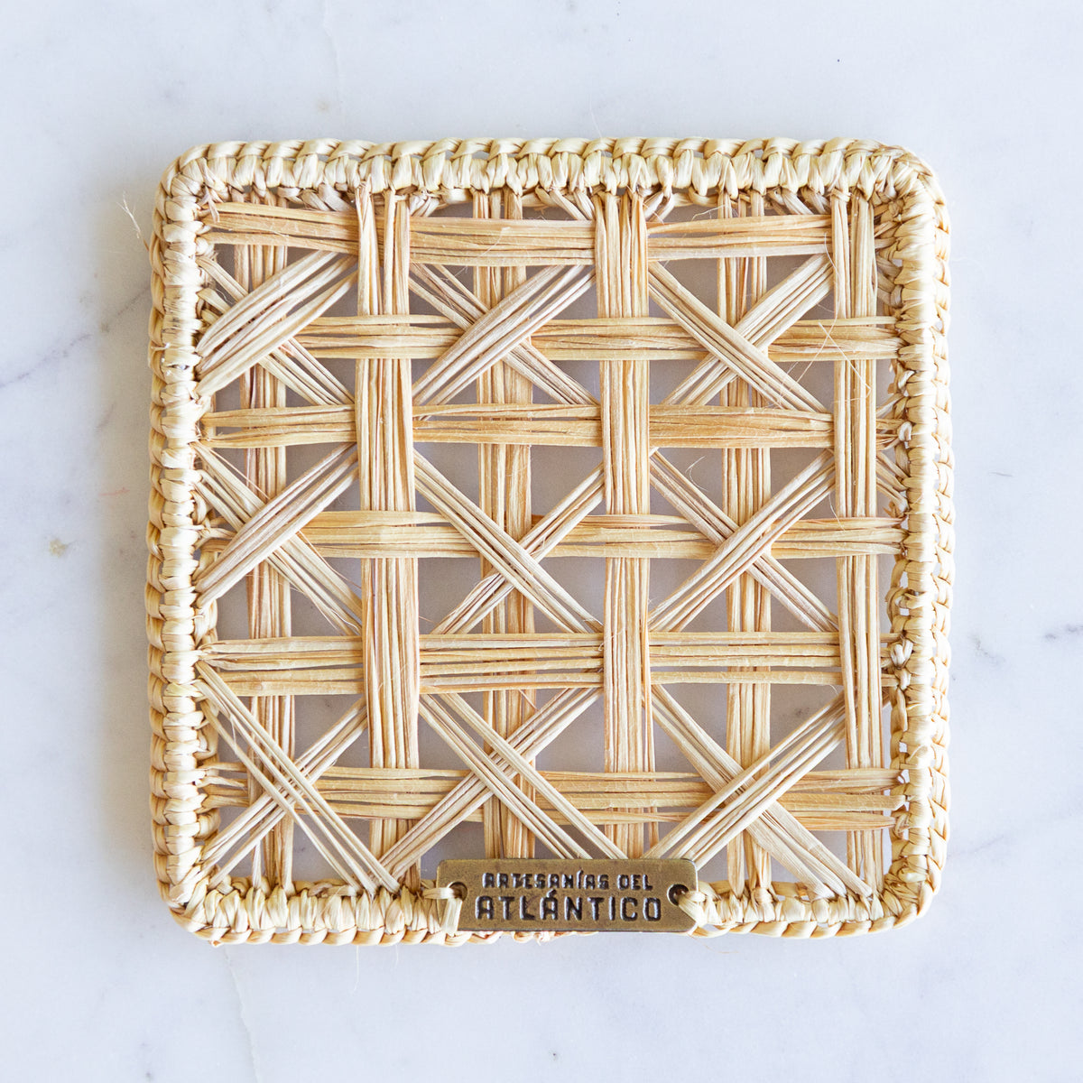 A square woven rattan decorative coaster with a thick braided border and a small metal brand plaque at the bottom, photographed from above on a light marble surface.