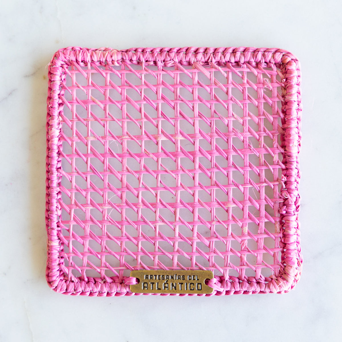 A square pink woven rattan coaster with a thick braided border and a small brass brand plaque at the bottom, photographed from above on a light marble surface.