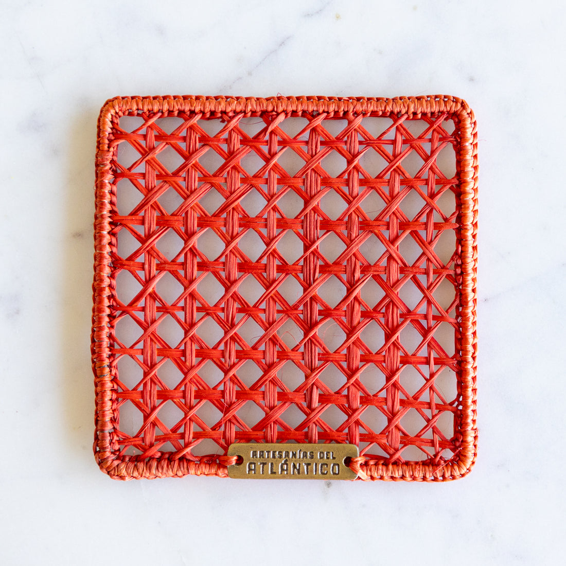 A square woven rattan coaster in a vibrant orange-red hue. The coaster is photographed from directly above on a white marble surface.