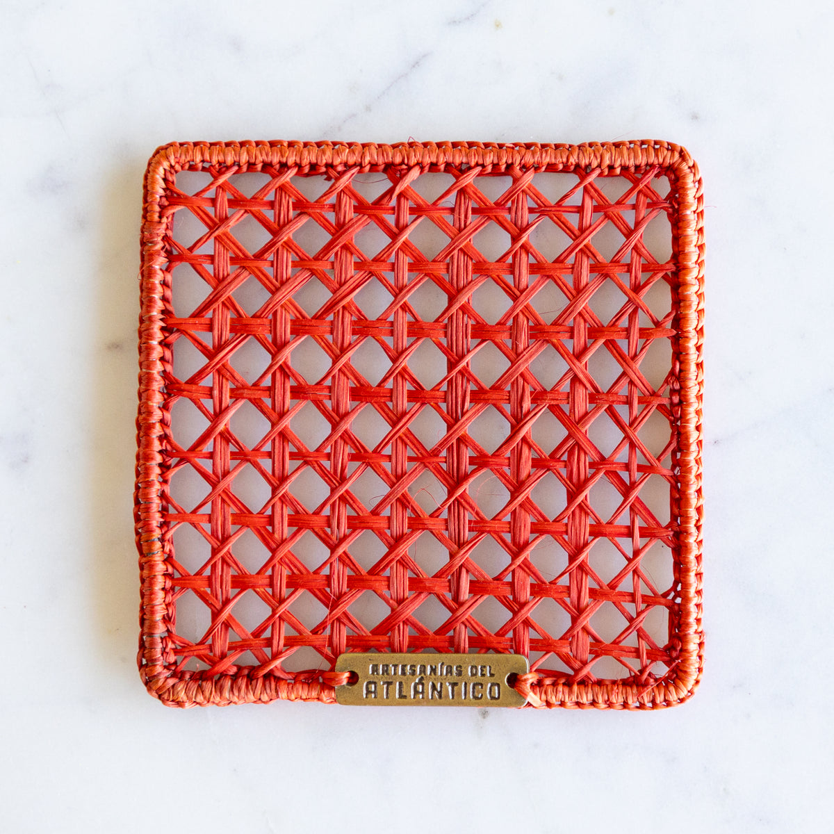 A square woven rattan coaster in a vibrant orange-red hue. The coaster is photographed from directly above on a white marble surface.