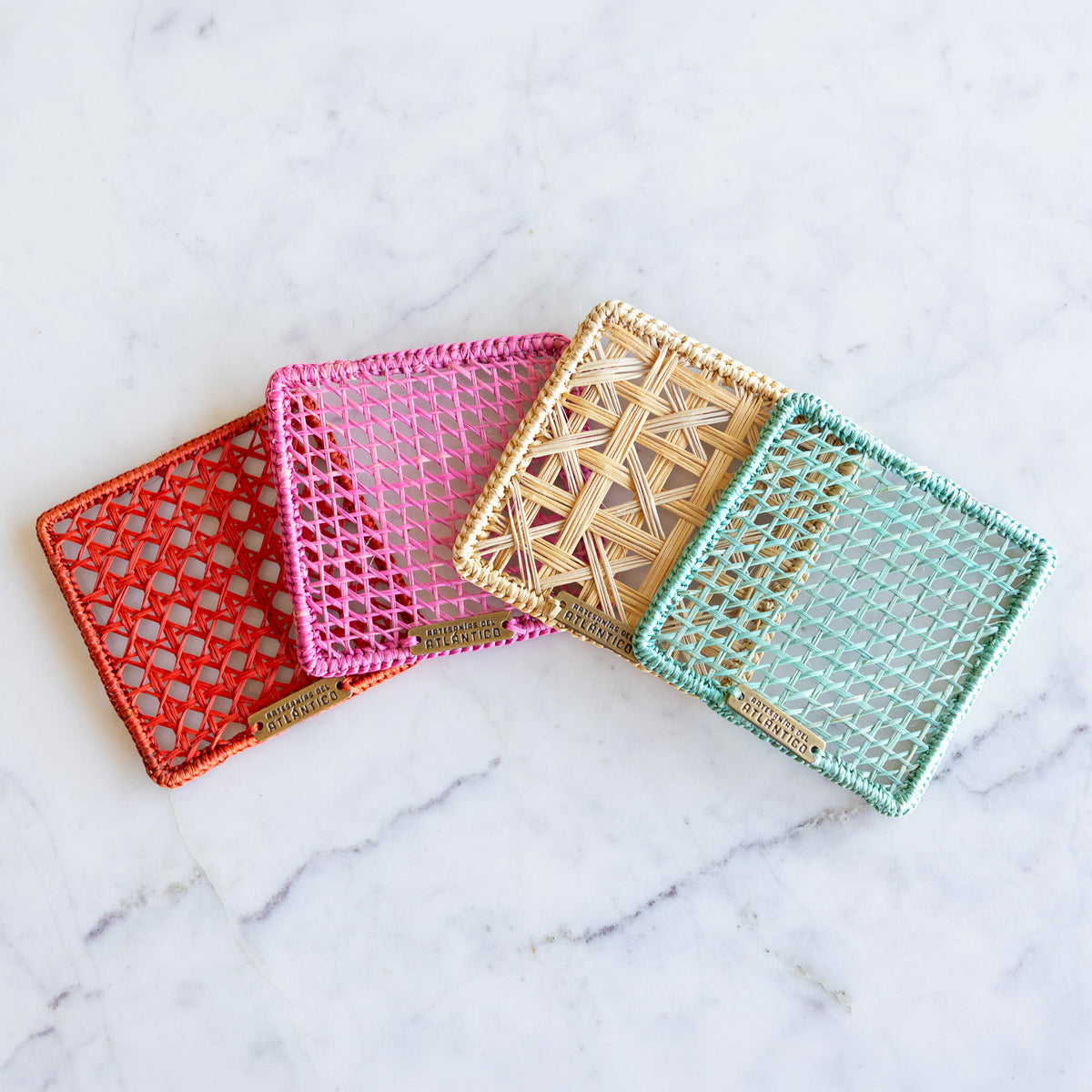 Four woven rectangular card coasters arranged in a fan on a white marble surface. The cases are shown in red, pink, beige, and mint green, each with an open lattice weave and a small gold-tone brand tag at the bottom edge.