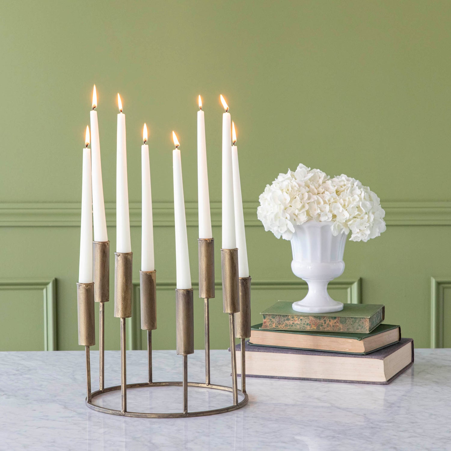 A short, iron taper candle holder with eight lit candles, on a table with flowers stacked on top of various sized books.