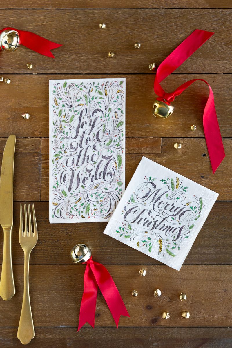Two napkins on a wooden table with holiday decorations and gold flatware, the guest napkin says "Joy to the World" and the cocktail napkin says "Merry Christmas". 