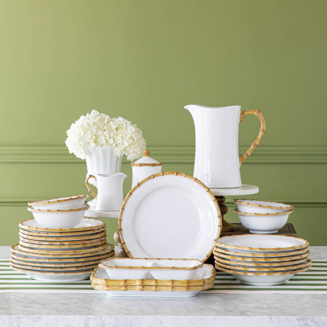 Juliska Bamboo dinnerware and serveware stacked together and on top of risers, with a vase of flowers, on a marble table.