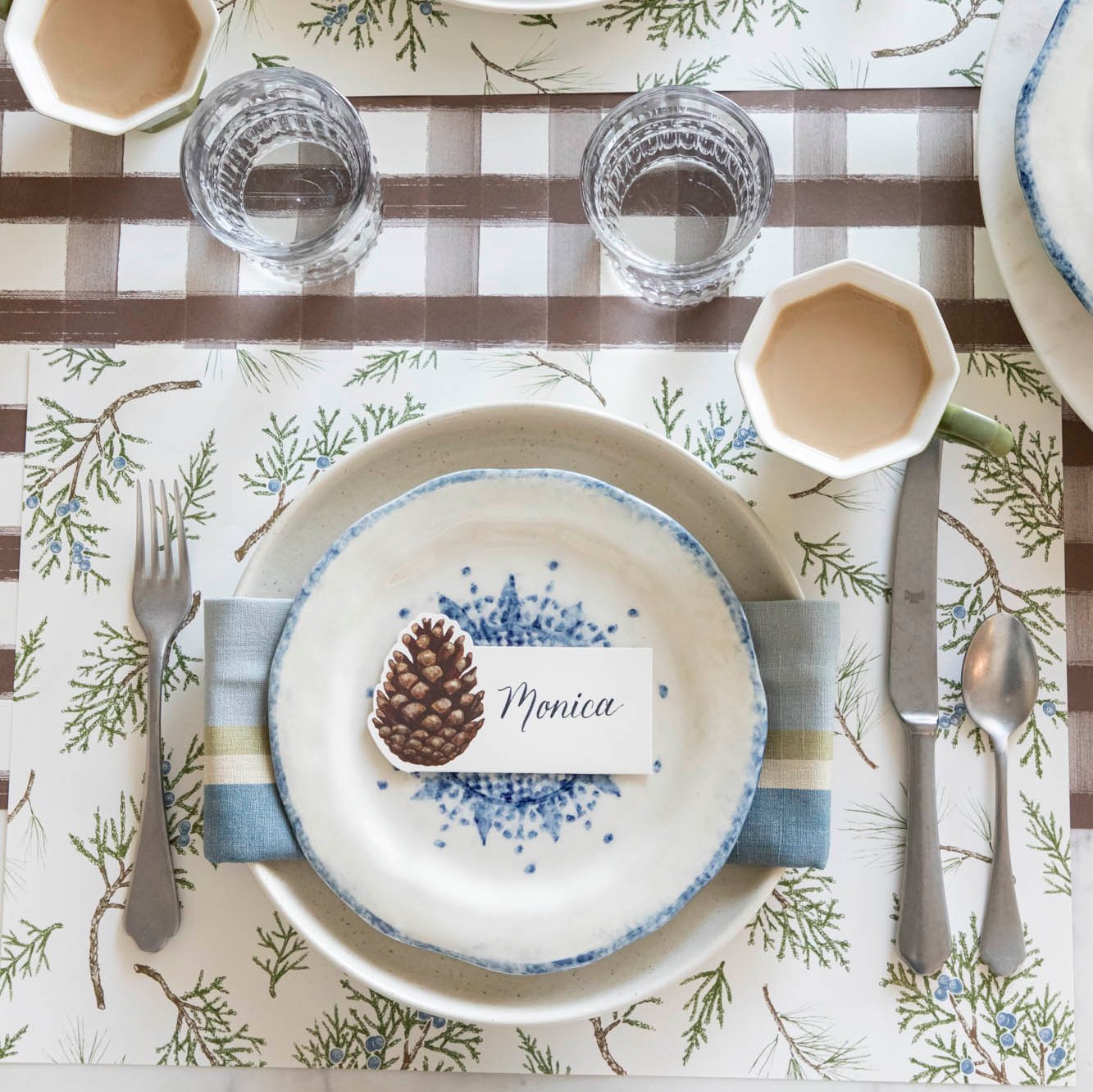 An elegant winter themed table setting with a Pinecone Place Card with &quot;Monica&quot; written on it, on top of dinner plates and a Juniper Sprigs Placemat underneath.