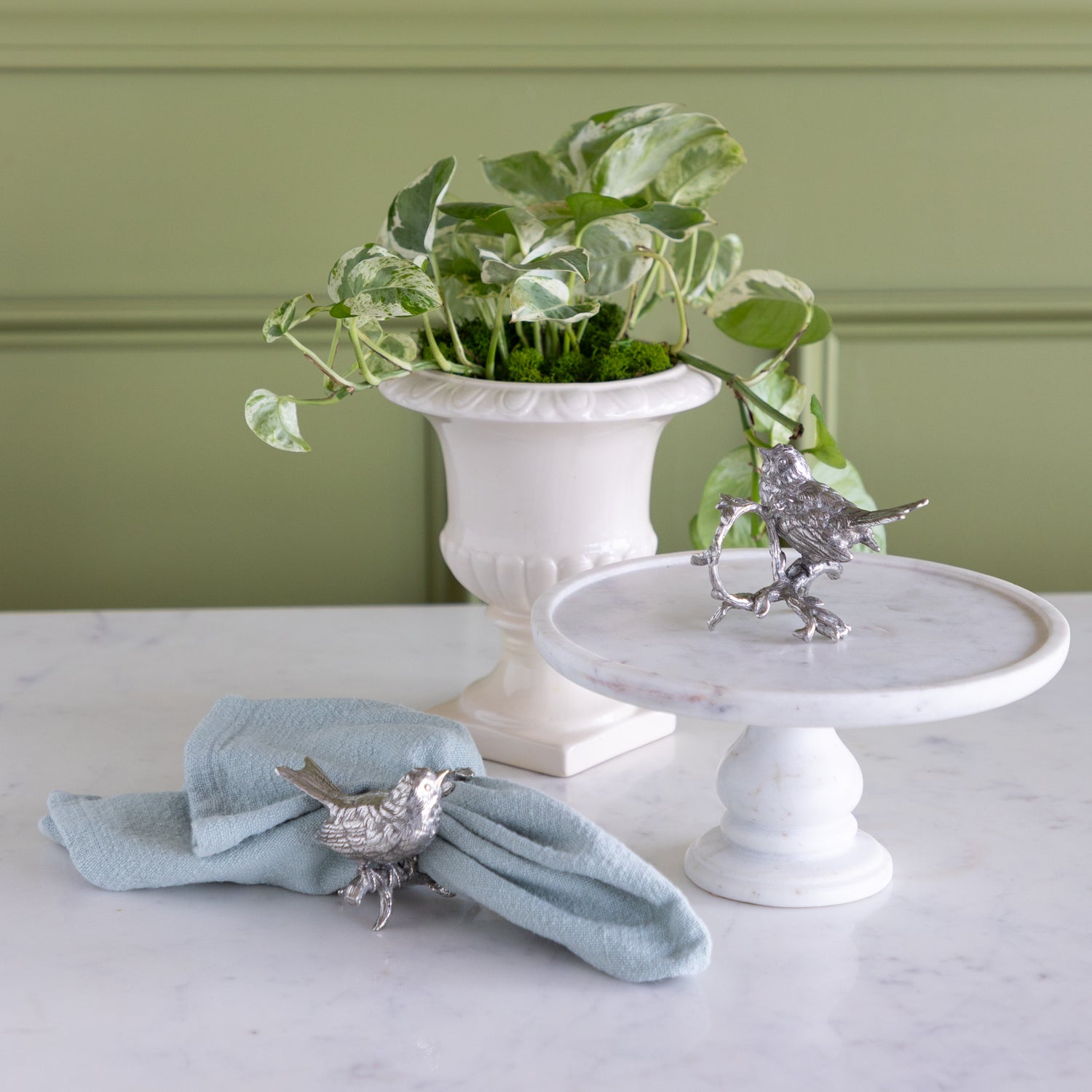 Decorative silver bird napkin ring on a light blue napkin and white pedestal with a plant in the background.