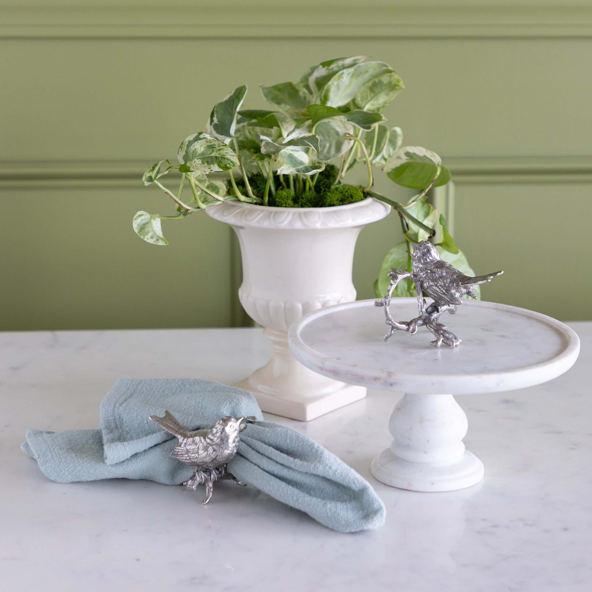Decorative silver bird napkin ring on a light blue napkin and white pedestal with a plant in the background.