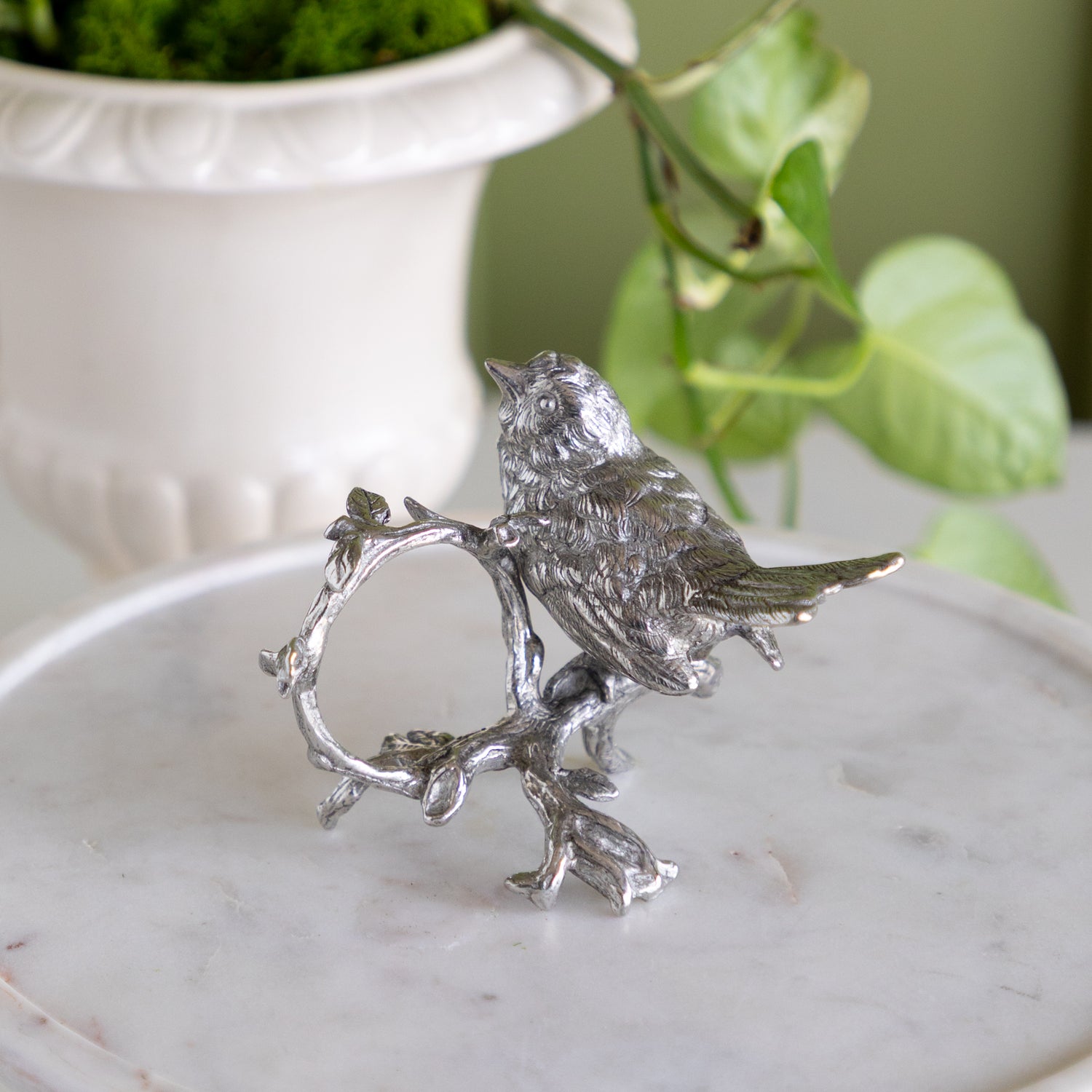 Decorative silver bird napkin ring on a white pedestal with a plant in the background.