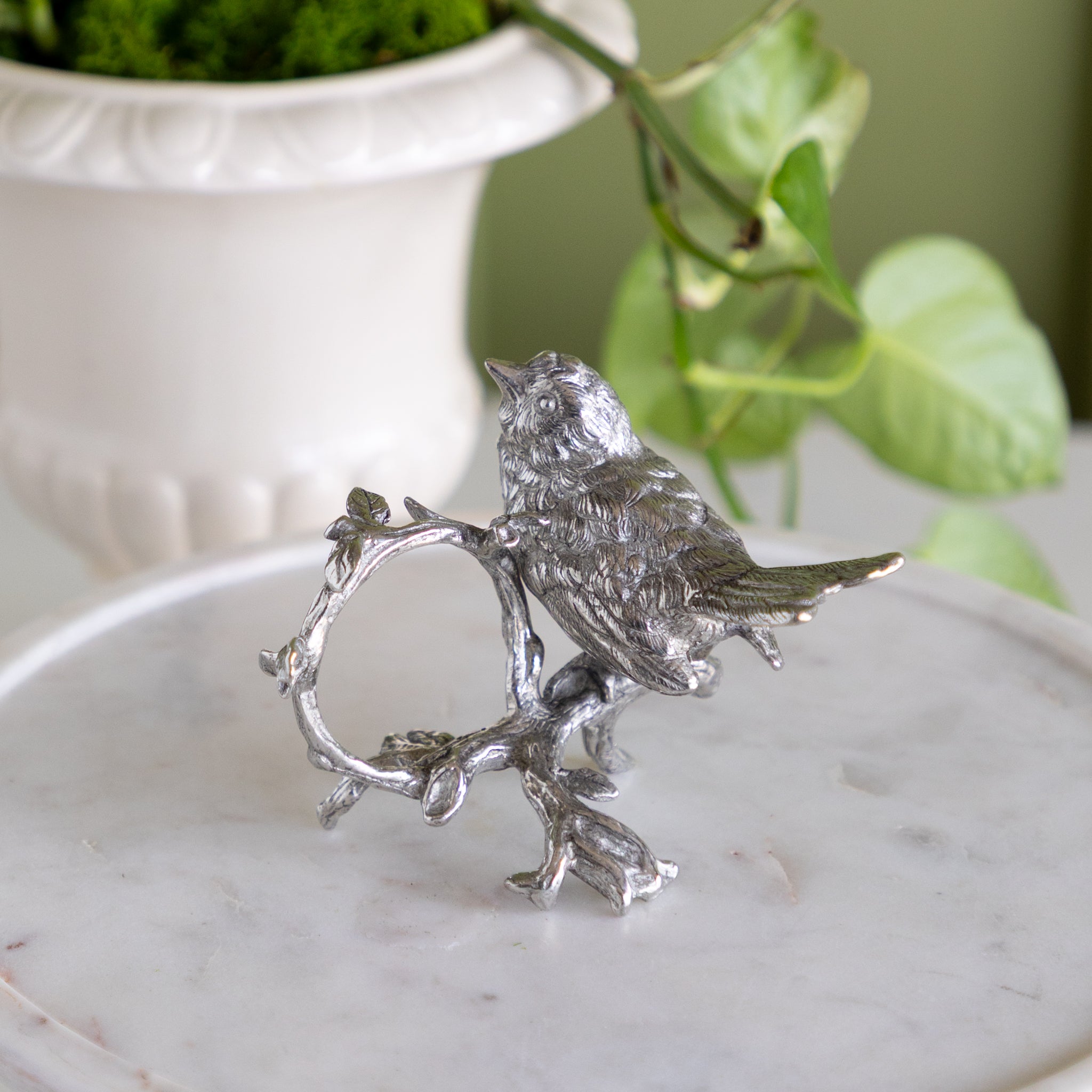 Decorative silver bird napkin ring on a white pedestal with a plant in the background.