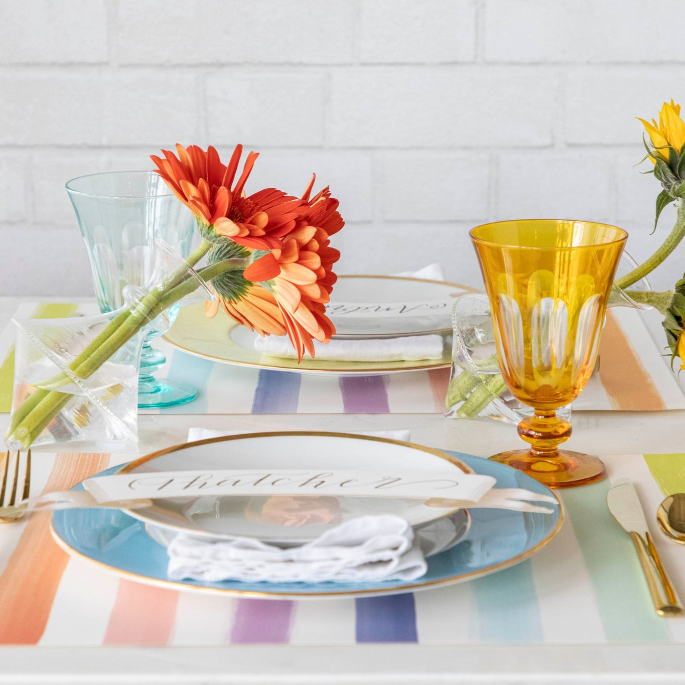 Colorful table setting with the Sorbet Painted Stripe Placemat, plates with the Gold Banner Table Accent atop, gold flatware and vases of flowers.