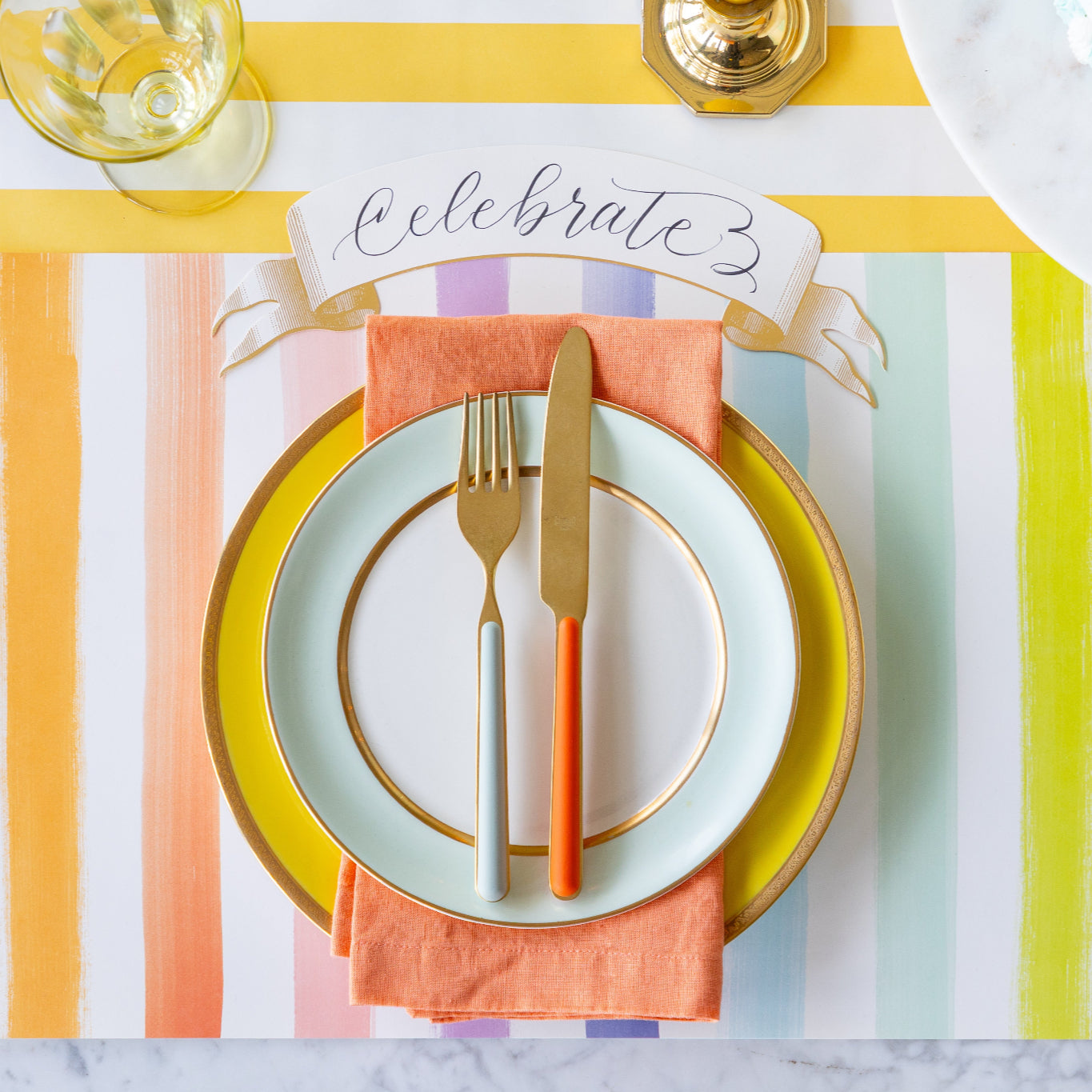 Colorful table setting with the Sorbet Painted Stripe Placemat, colorful plates and cutlery atop, and Classic Gold Banner Table Accent on the Marigold Classic Stripe Runner.