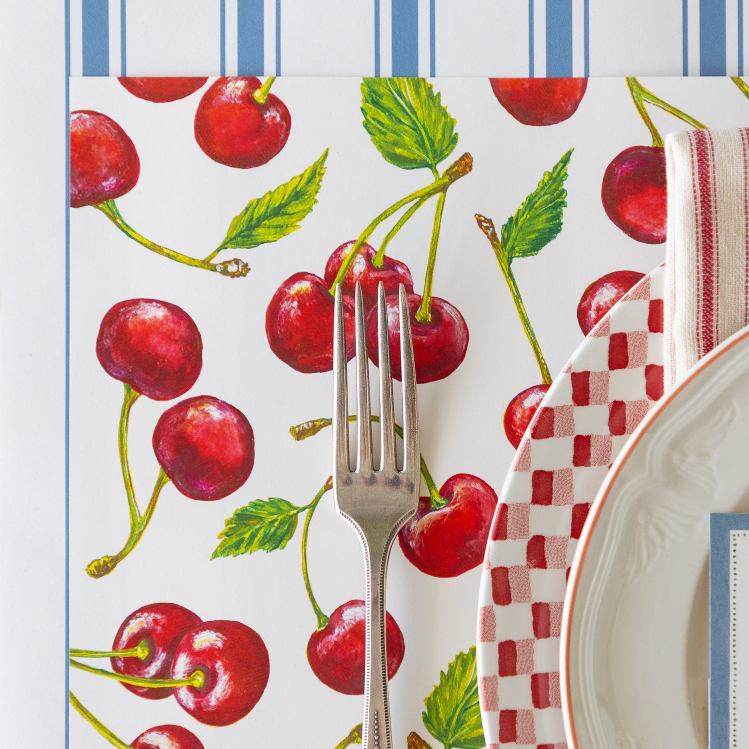 Placemat with cherry design, fork, plate, and name card on the Cornflower Blue Linen Stripe Runner.