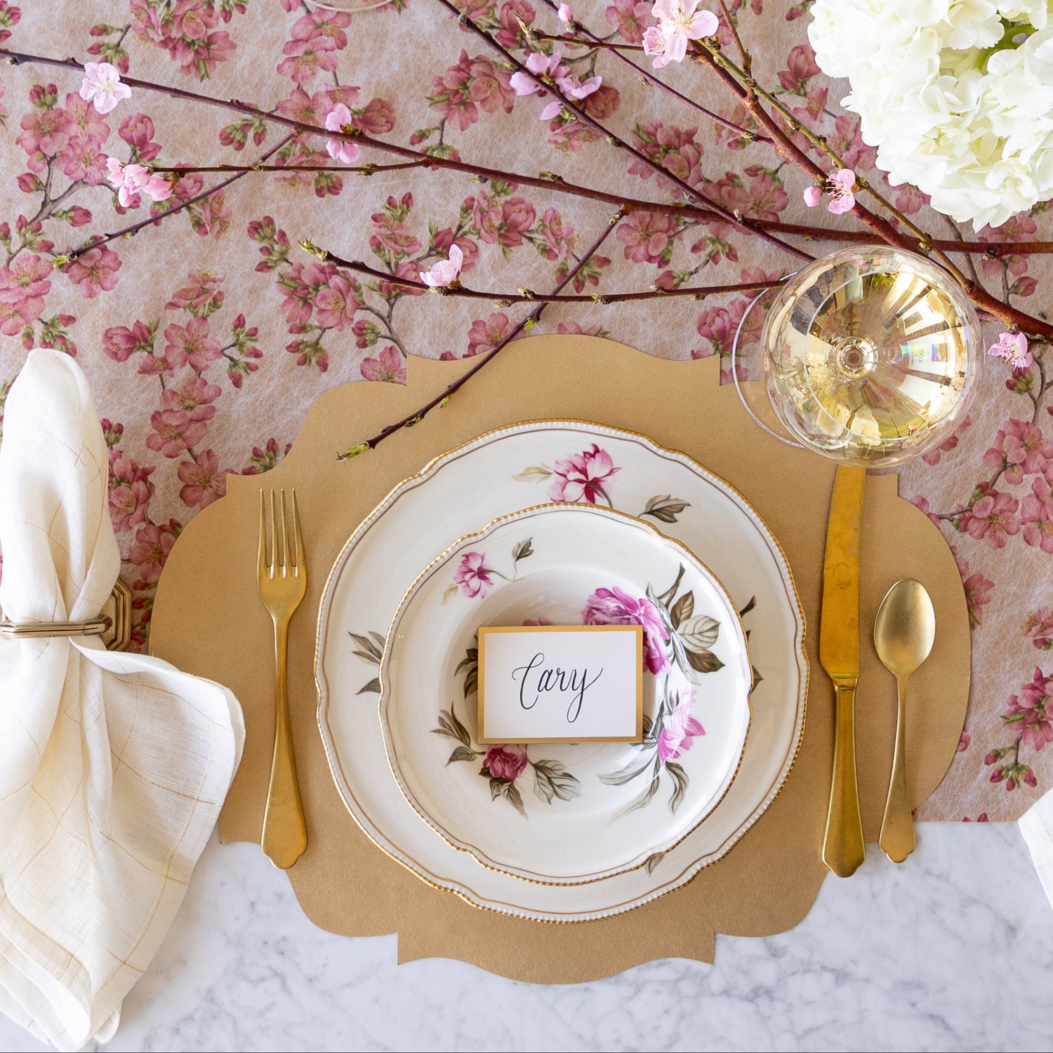 An elegant table setting with flowers and a cherry blossom branch, featuring the Die-cut Gold French Frame Placemat under vintage floral dinnerware with the Gold Frame Place Card reading &quot;Cary&quot; atop, gold flatware, a glass of wine and Cherry Blossom Runner underneath.