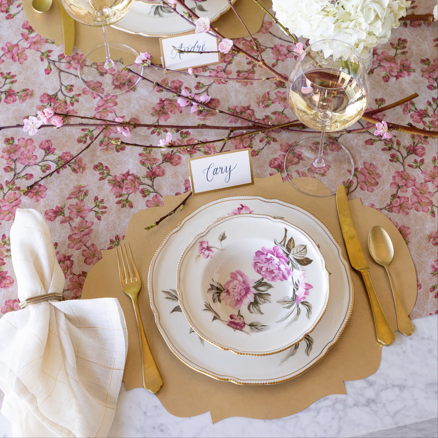 An elegant table setting with flowers and a cherry blossom branch, featuring the Die-cut Gold French Frame Placemat under vintage floral dinnerware, the Gold Frame Place Card reading &quot;Cary&quot;, gold flatware, a glass of wine and Cherry Blossom Runner underneath.