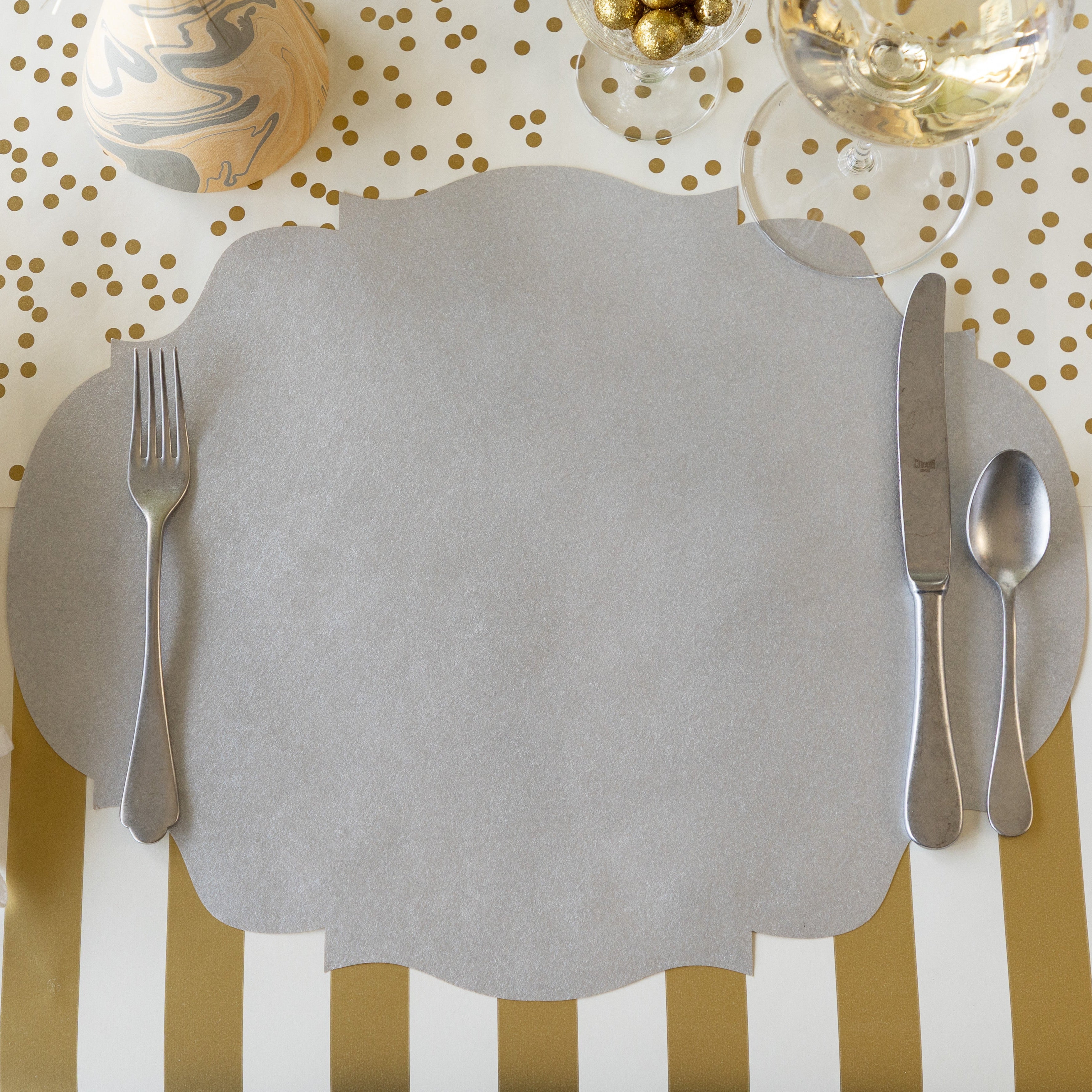 The Die-cut Silver French Frame Placemat with vintage flatware on top, a glass of wine, marbled party hat, Classic Gold Stripe Runner rolled out vertically and Gold Confetti Runner rolled out horizontally underneath.