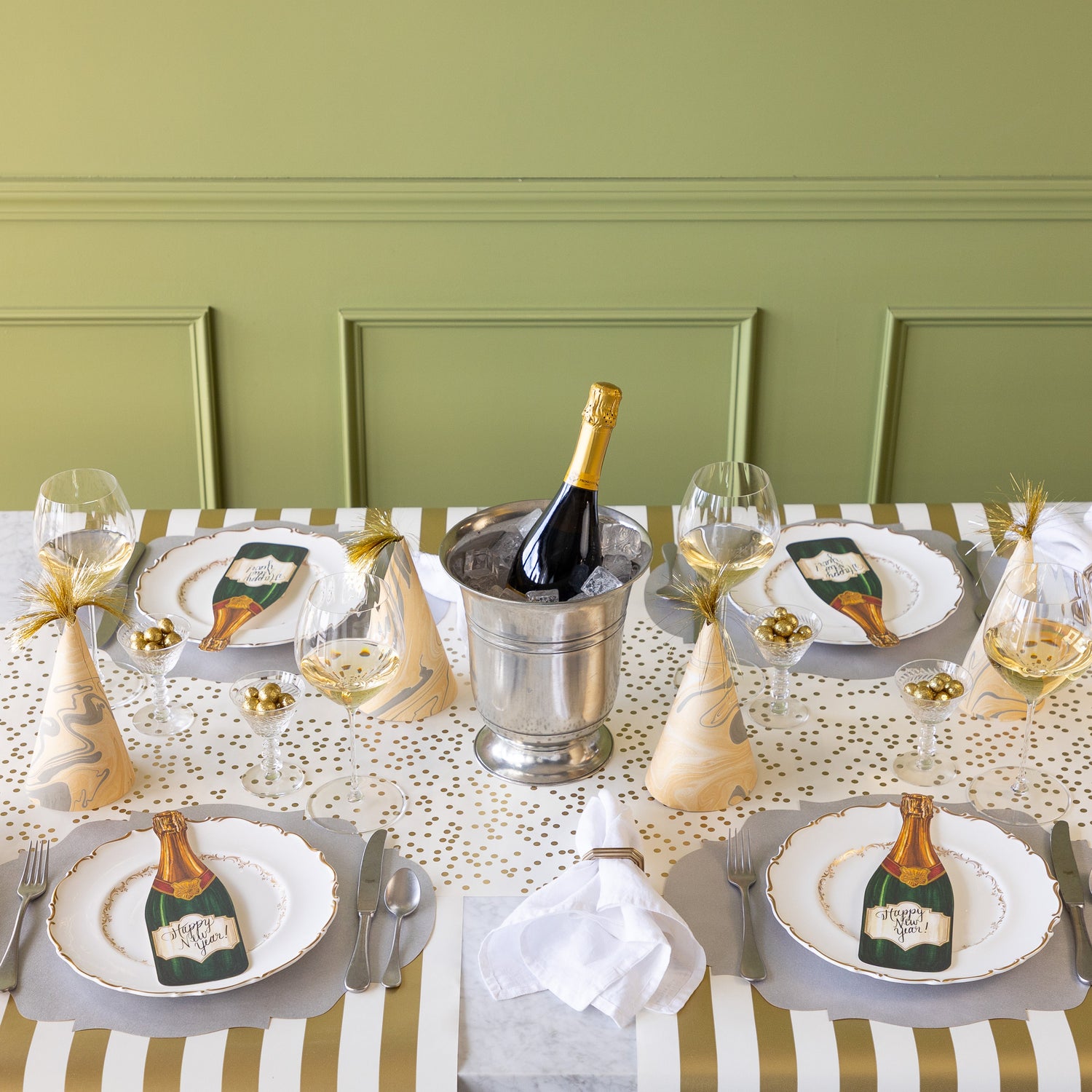 A table for four is set with champagne in an ice bucket, marbled party hats, Die-cut Silver French Frame Placemat under a dinner plate with the Champagne Table Accents atop, glasses of wine, cups of gold glitter balls, Classic Gold Stripe Runner rolled out vertically and Gold Confetti Runner rolled out horizontally on the table.
