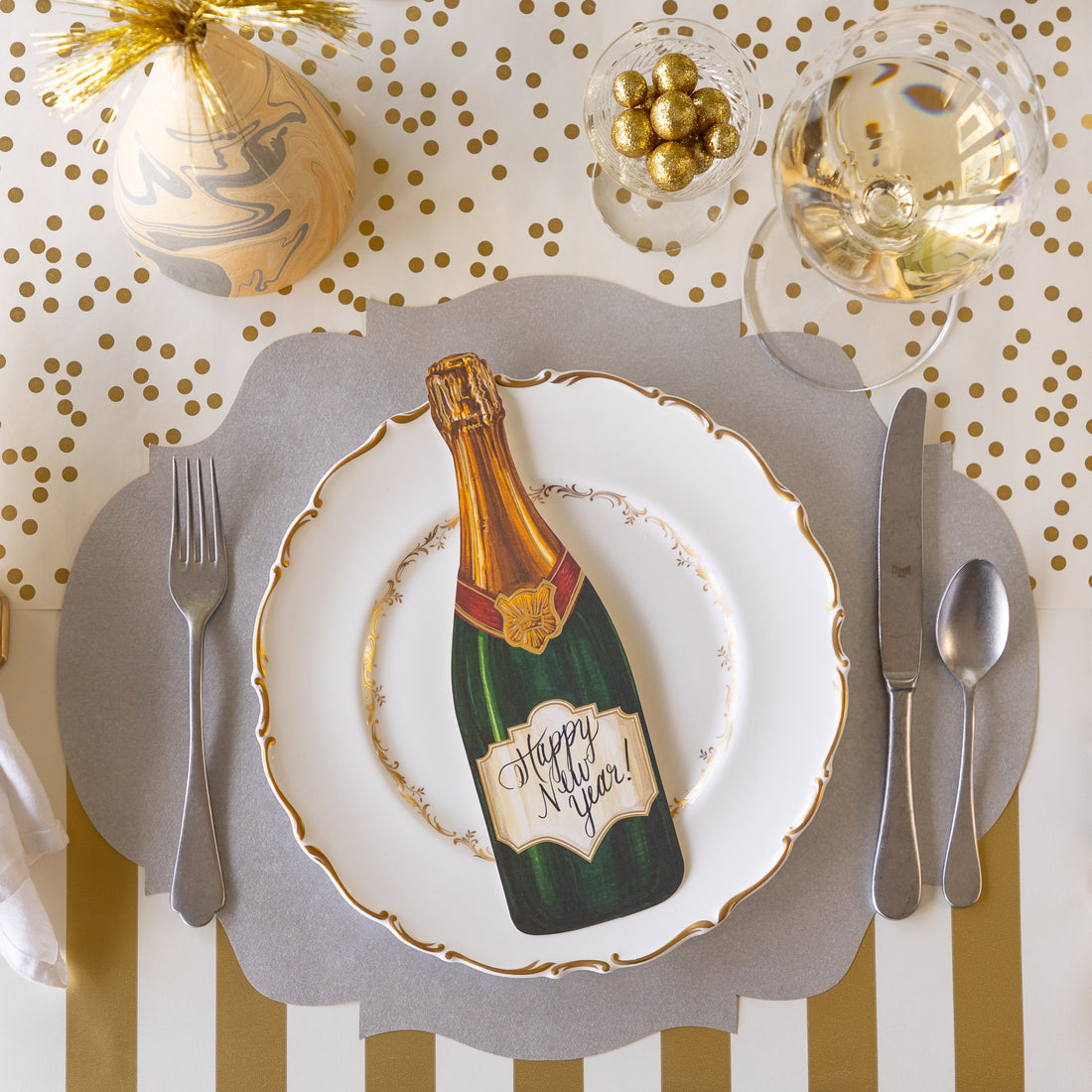 An elegant place setting featuring the Die-cut Silver French Frame Placemat, a gold rimmed dinner plate with the Champagne Table Accent reading "Happy New Year!" atop, a marbled party hat, glass of wine, Classic Gold Stripe Runner rolled out vertically and Gold Confetti Runner rolled out horizontally underneath.