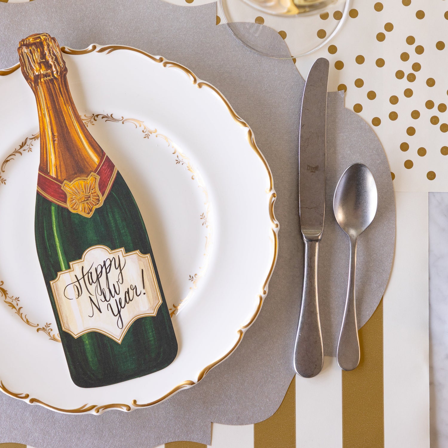 An elegant place setting featuring the Die-cut Silver French Frame Placemat under a gold rimmed dinner plate with the Champagne Table Accent atop, vintage silver flatware, Classic Gold Stripe Runner rolled out vertically underneath and Gold Confetti Runner rolled out horizontally.