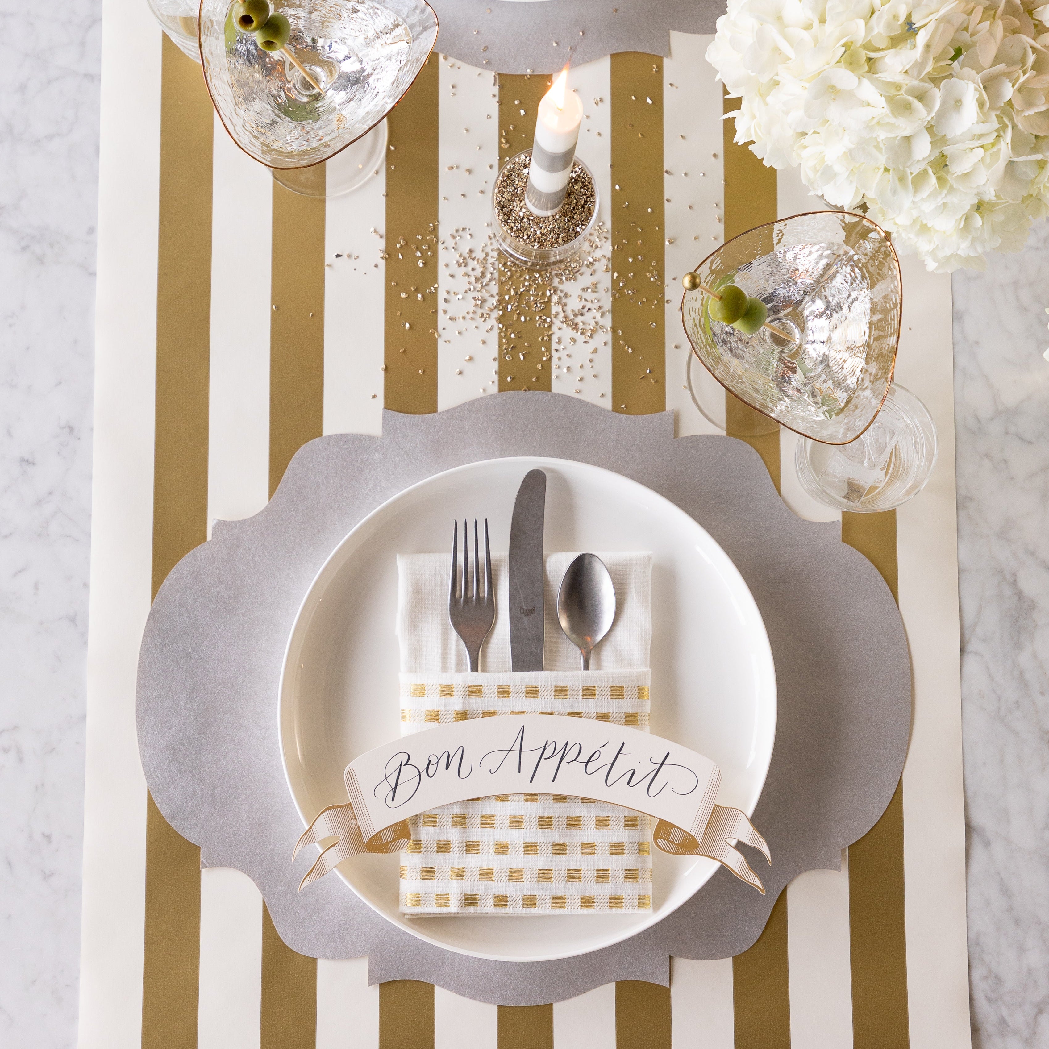 A place setting featuring the Die-cut Silver French Frame Placemat with silverware inside of a folded napkin and Classic Gold Banner Table Accent reading &quot;Bon Appetit&quot; on a dinner plate, a martini glass with olives, a lit candle, flowers and the Gold Classic Stripe Runner underneath.