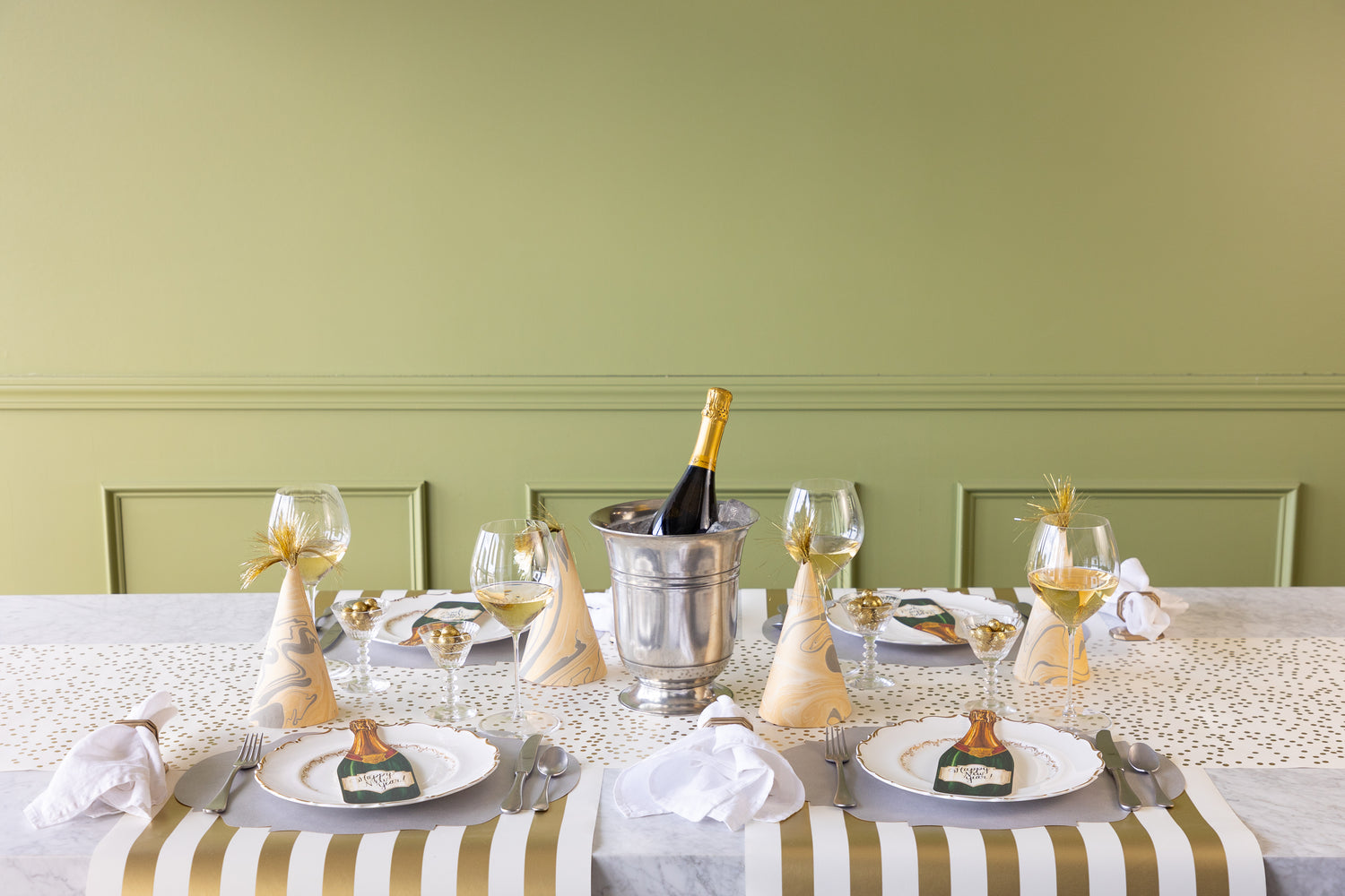 A festive table with the confetti runner on it and party hats at 4 place settings.  White plates and a bottle of champagne are also on the table. 