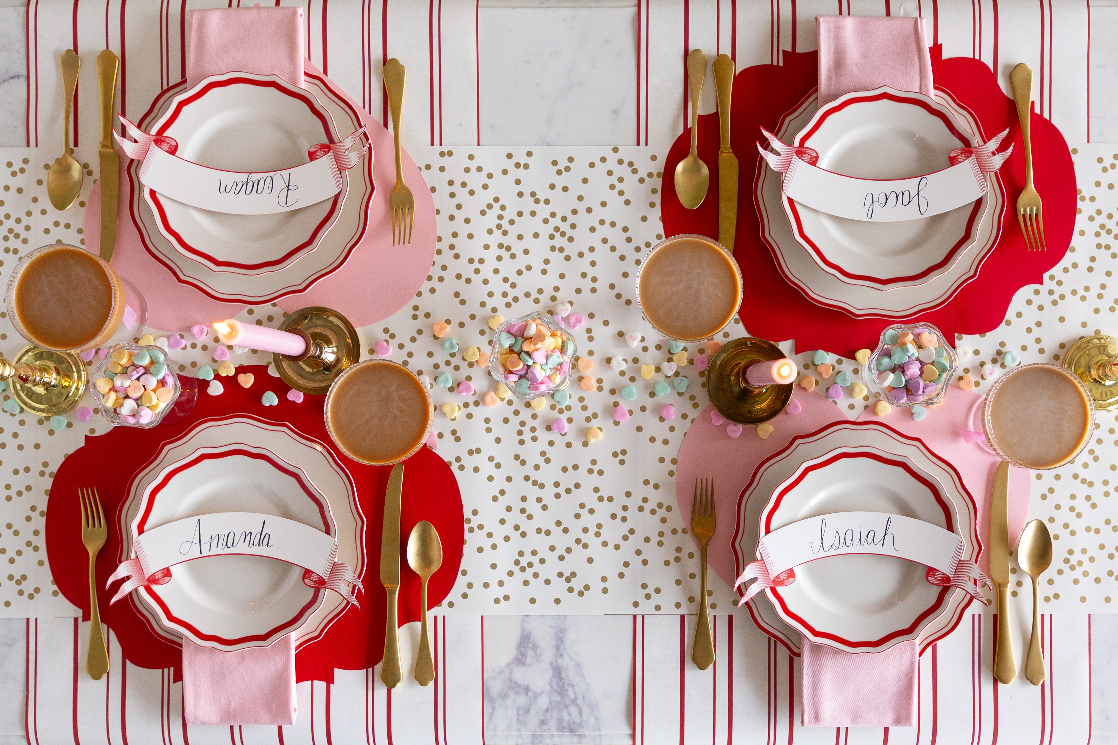 A Valentine's table set with Die-cut Pink Heart and Red French Frame Placemats, Red Icing on the Cake Dinnerware, Classic Red Banner Table accents, candles, candy and gold flatware on the Gold Confetti and Antique Red Stripe Runners. 