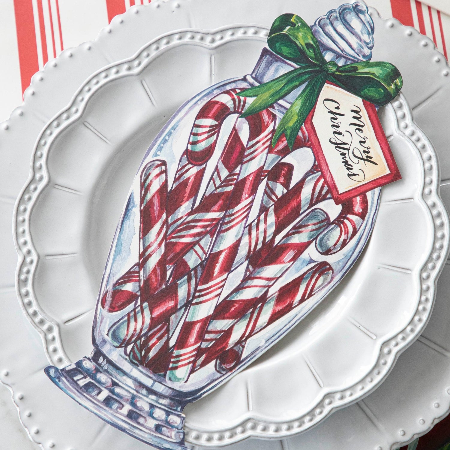 A close up of the Candy Cane Jar Table Accent with &quot;Merry Christmas&quot; written on the tag, resting on white dinner plates.