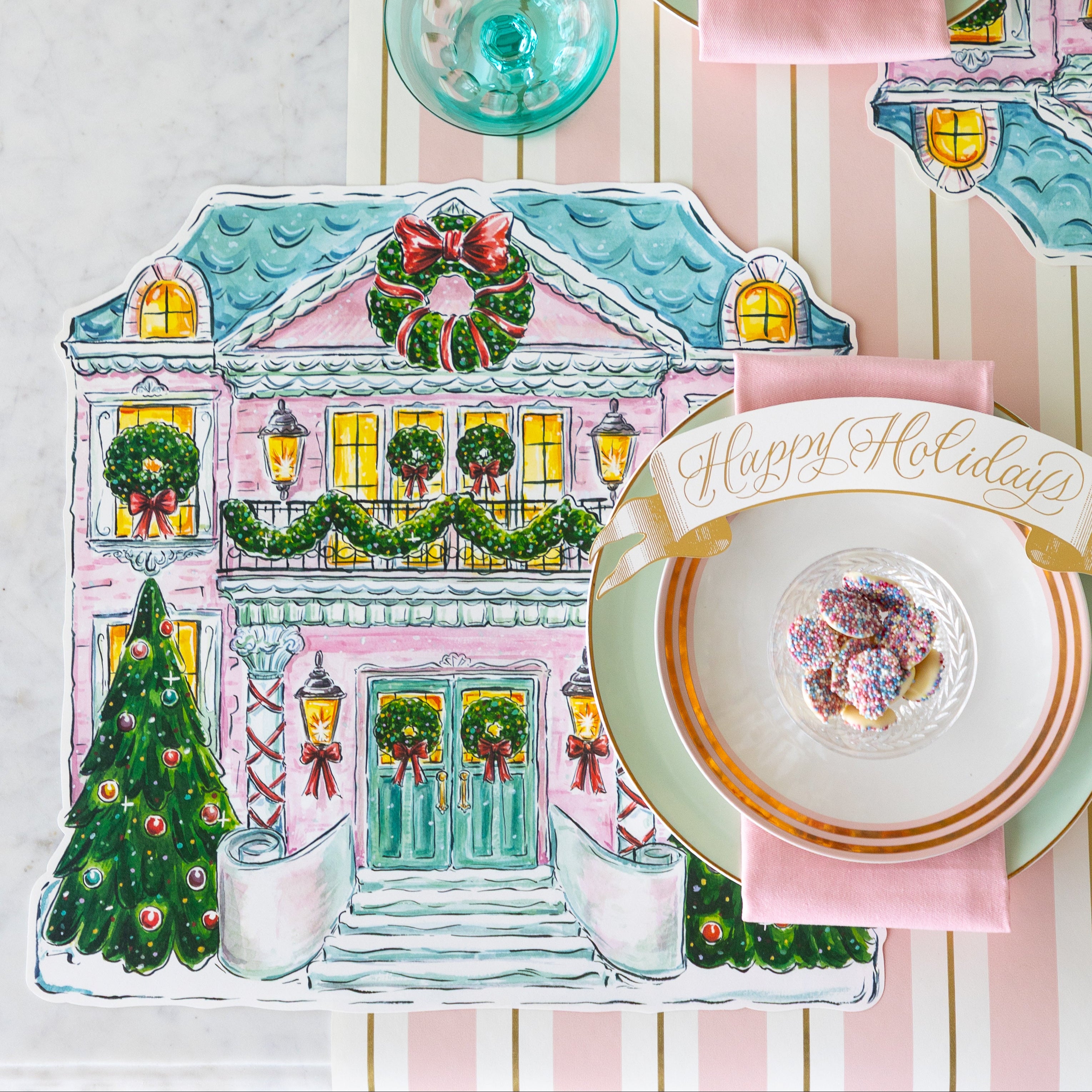 A place setting featuring the Die-cut Holiday Home Placemat, seafoam and gold-rimmed plates, a  pink napkin, bowl of white chocolate buttons and Happy Holidays Banner Table Accent atop, on the Pink &amp; Gold Awning Stripe Runner.
