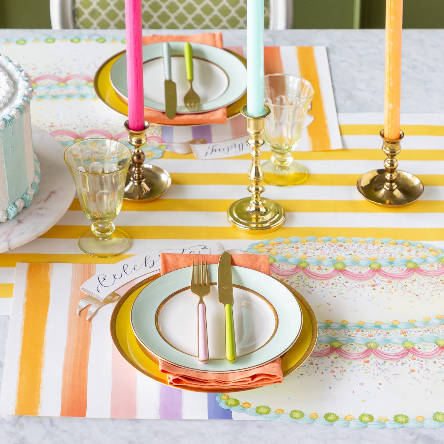 A colorful table setting featuring the Die-cut Birthday Cake Placemat overlapping the Sorbet Painted Stripe Placemat, colorful dinnerware and Classic Gold Banner Table Accent on the Marigold Classic Stripe Runner.