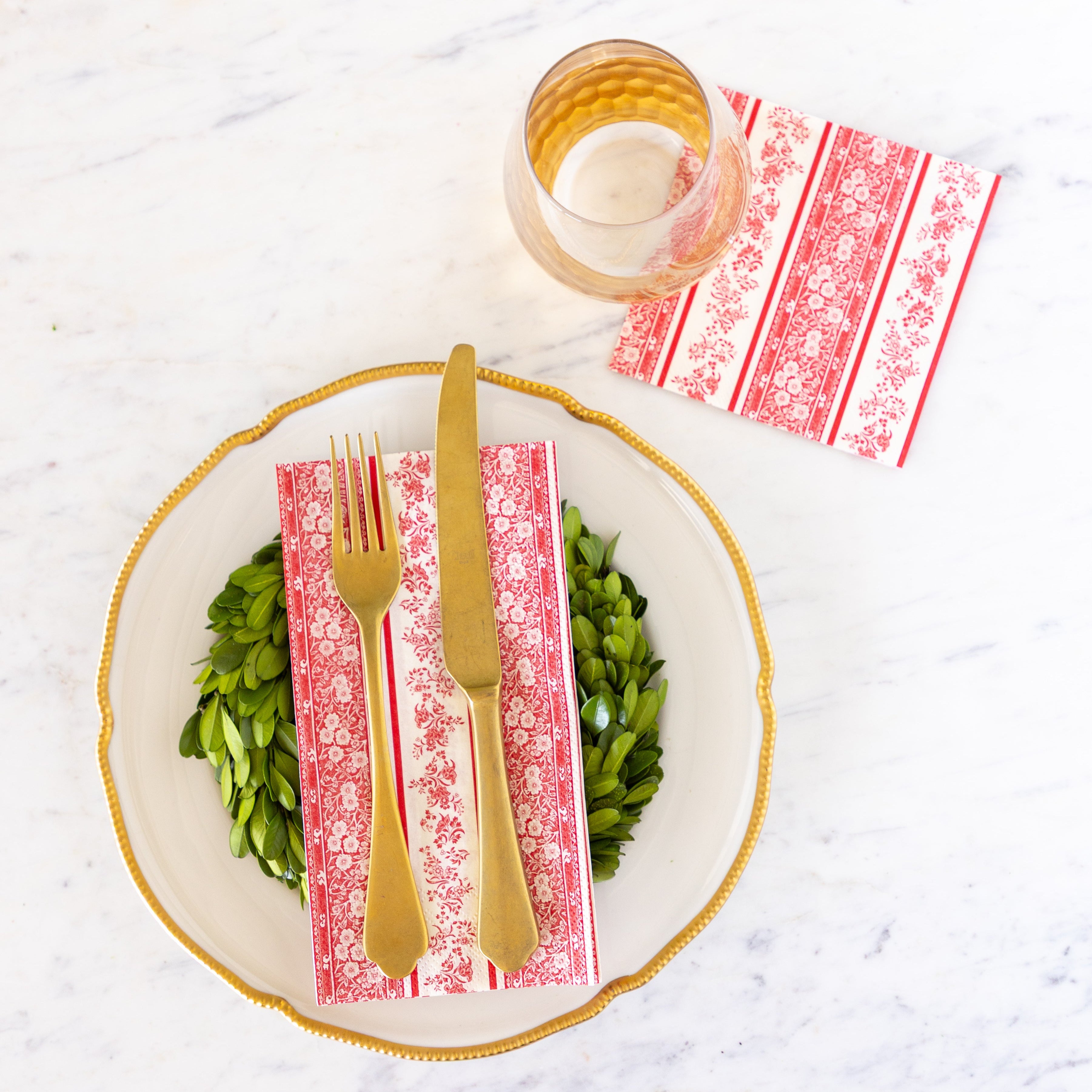 A plate with a boxwood wreath, Red Burleigh Stripe Guest Napkin and gold flatware on top, and a glass slightly on top of the Red Burleigh Stripe Cocktail Napkin.