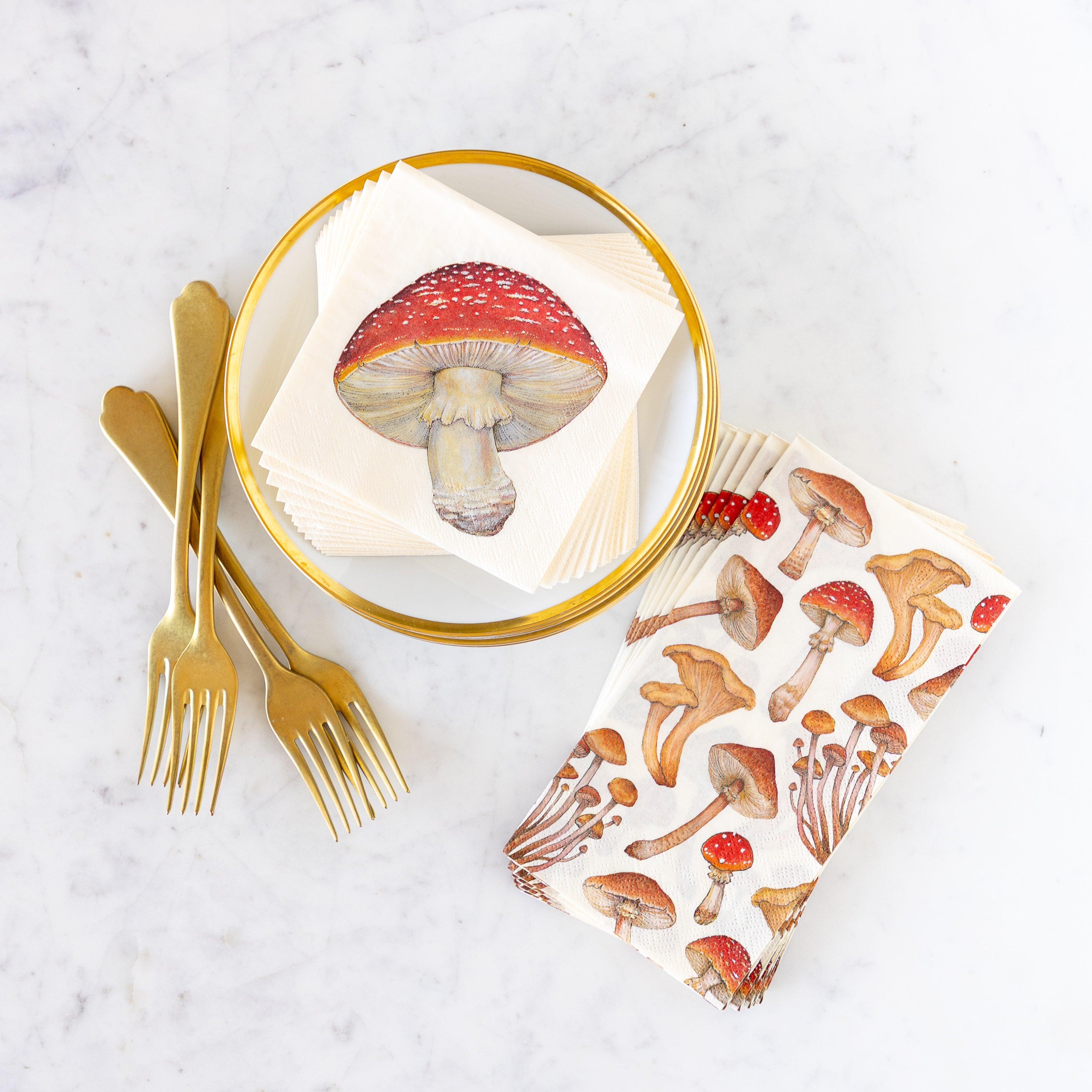 A fanned out stack of Mushroom Cocktail Napkins on a gold rimmed plate next to forks, and a stack of Mushroom Guest Napkins beside it.
