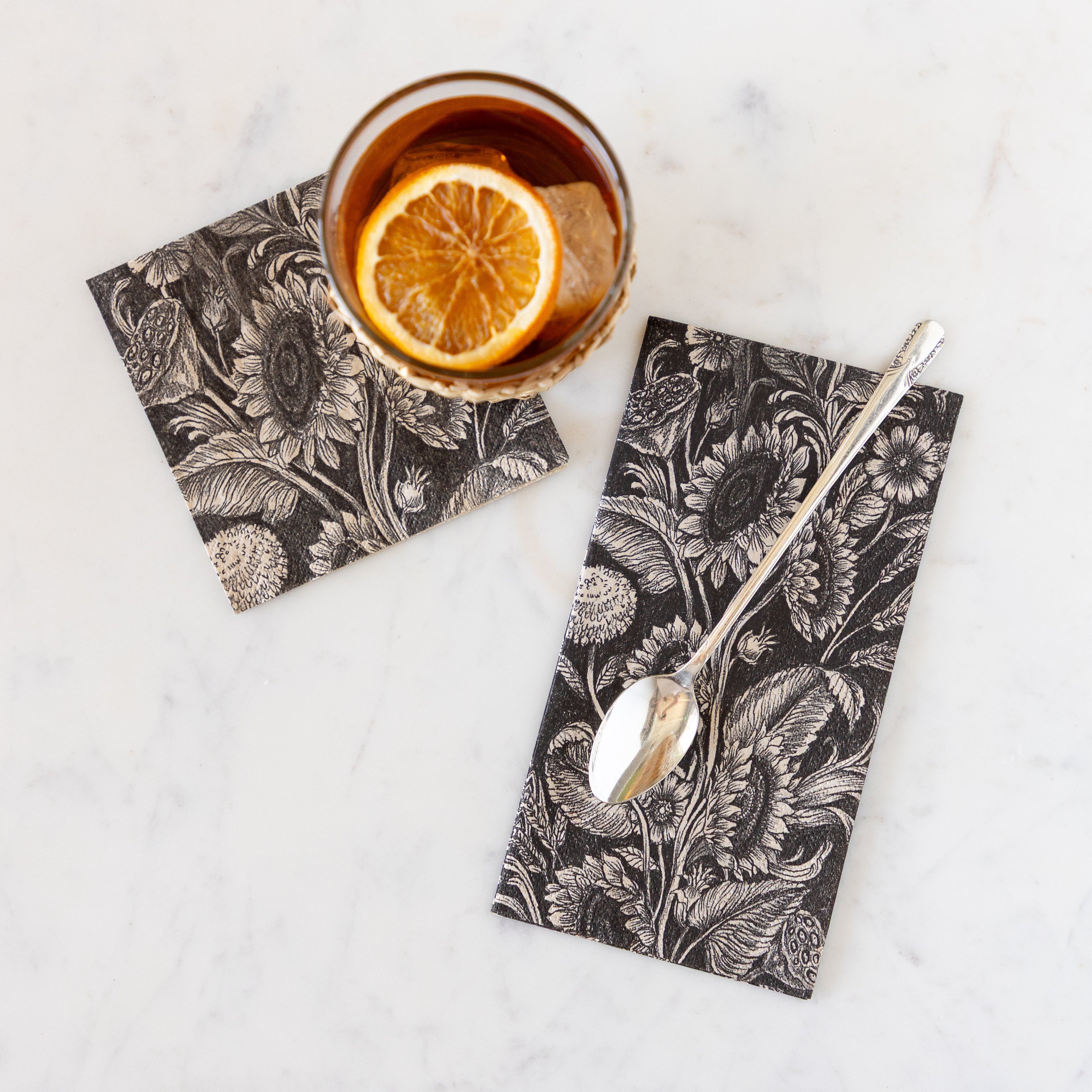 A glass of tea with an orange slice in it slightly on the Vintage Sunflower Cocktail napkin and a long handled tea spoon on the Vintage Sunflower Guest Napkin.