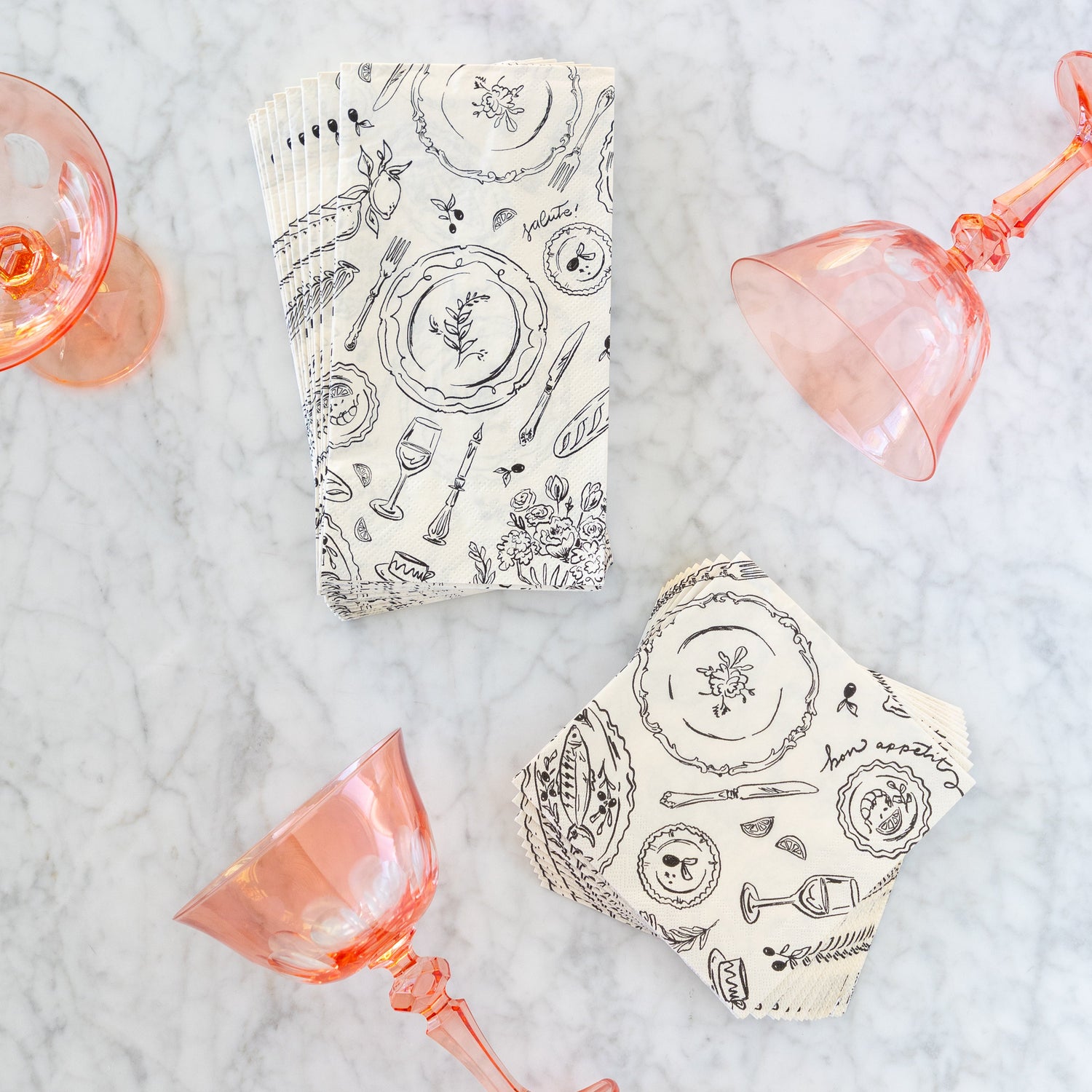 Dinner Party Guest and Cocktail Napkins surrounded by pink coupe glasses on a marble table.