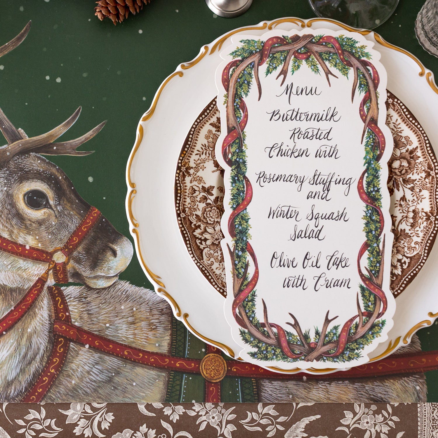 A festive place setting featuring the Antler Garland Table Menu resting on dinner plates, on the Holiday Reindeer Placemat.