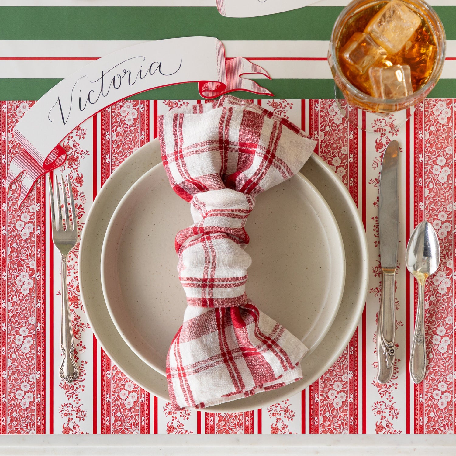 A place setting featuring the Red Burleigh Stripe Placemat, dinner plates with a knotted red and white checkered napkin atop, vintage silverware, a glass of tea, Red Classic Banner Table Accent reading &quot;Victoria&quot;, and Green &amp; Red Awning Stripe Runner underneath.