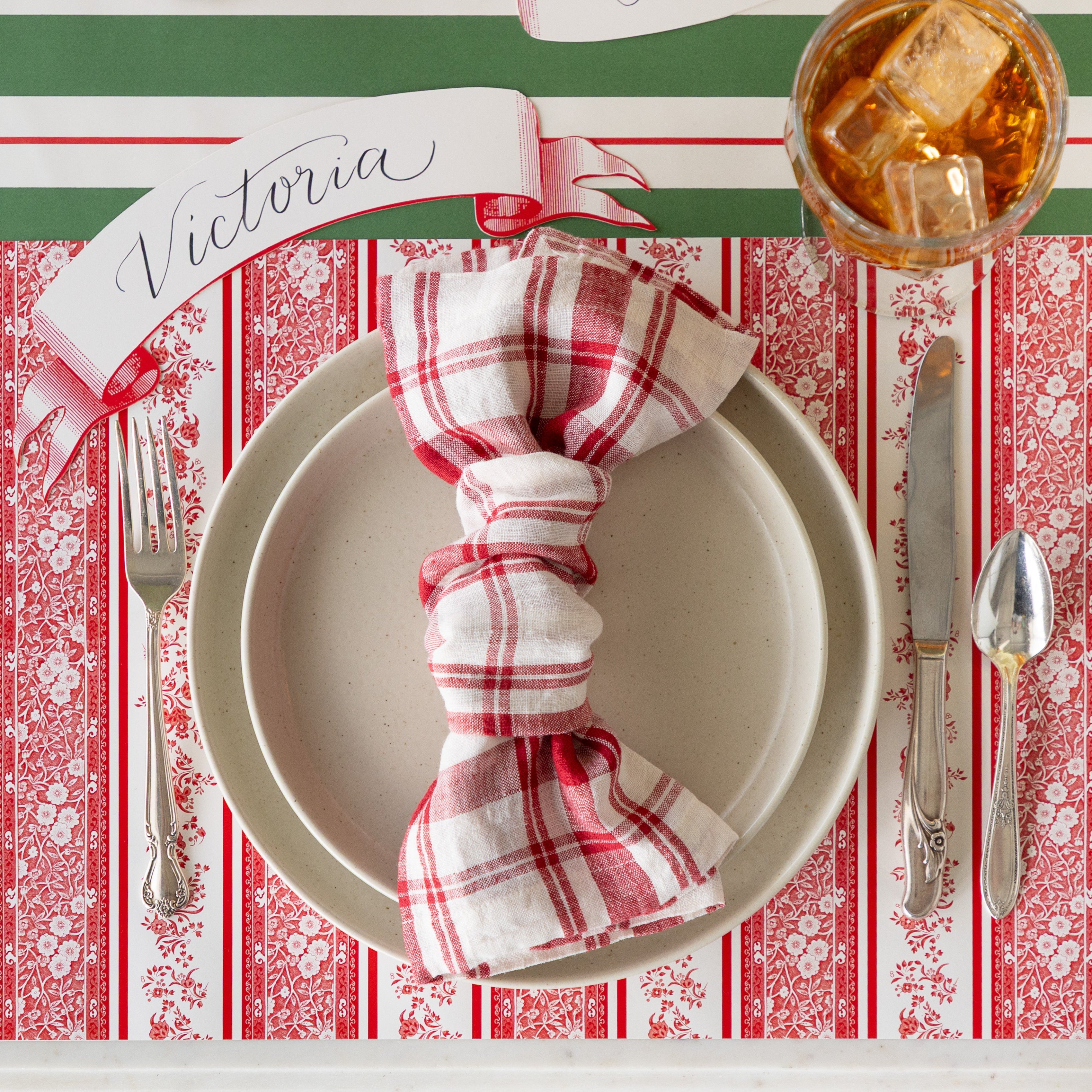 A place setting featuring the Red Burleigh Stripe Placemat, dinner plates with a knotted red and white checkered napkin atop, vintage silverware, a glass of tea, Red Classic Banner Table Accent reading &quot;Victoria&quot;, and Green &amp; Red Awning Stripe Runner underneath.