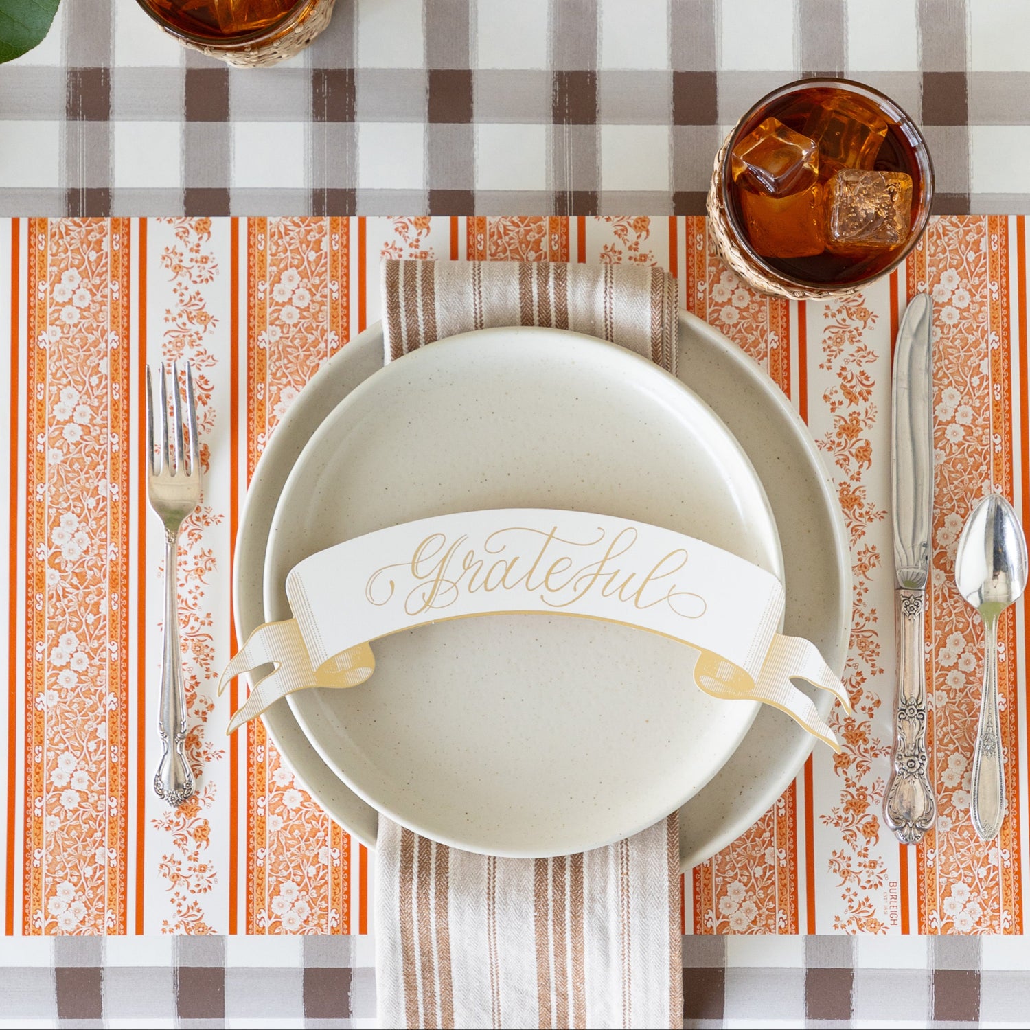 A place setting featuring the Pumpkin Burleigh Stripe Placemat with vintage silverware, vanilla dinnerware, a tan striped napkin and Grateful Banner Table Accent atop, on the Brown Painted Check Runner.