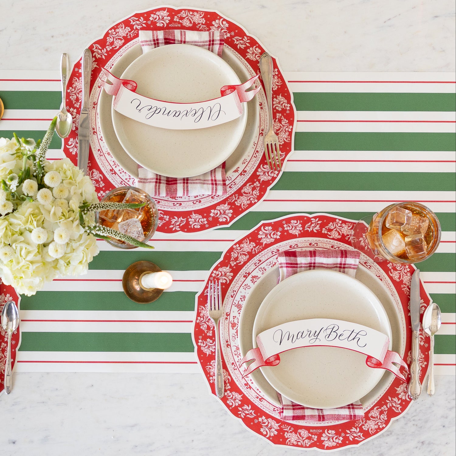 A table setting with a floral centerpiece and a lit candle, featuring the Die-cut Red Regal Peacock Placemat, off-white dinner plates with a red-and-white checkered napkin in between and Classic Red Banner Table Accent atop, vintage flatware, glasses of tea, and Green &amp; Red Awning Stripe Runner underneath.