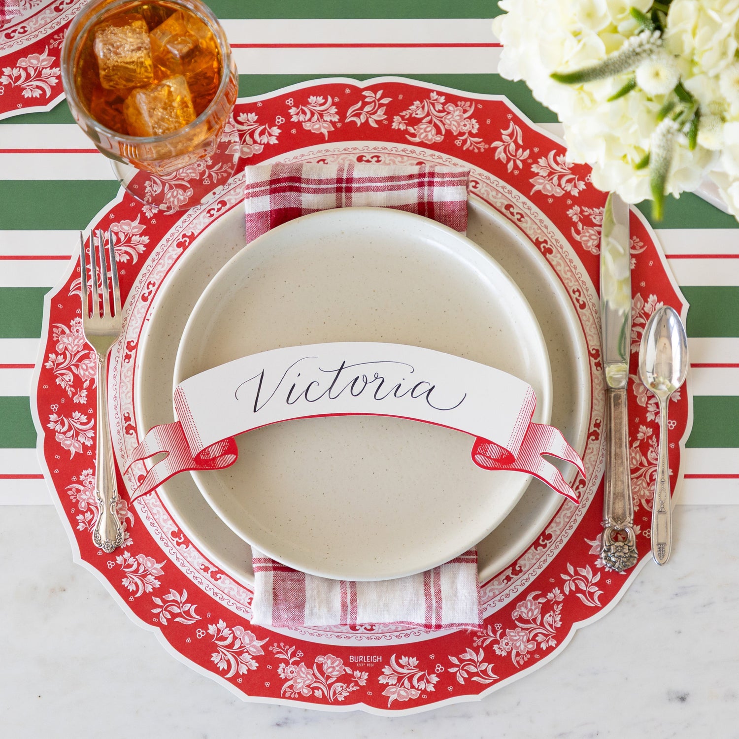 A place setting featuring the Die-cut Red Regal Peacock Placemat, off-white dinner plates with a red-and-white checkered napkin in between and Classic Red Banner Table Accent reading &quot;Victoria&quot; atop, vintage flatware, a glass of tea, and Green &amp; Red Awning Stripe Runner underneath.