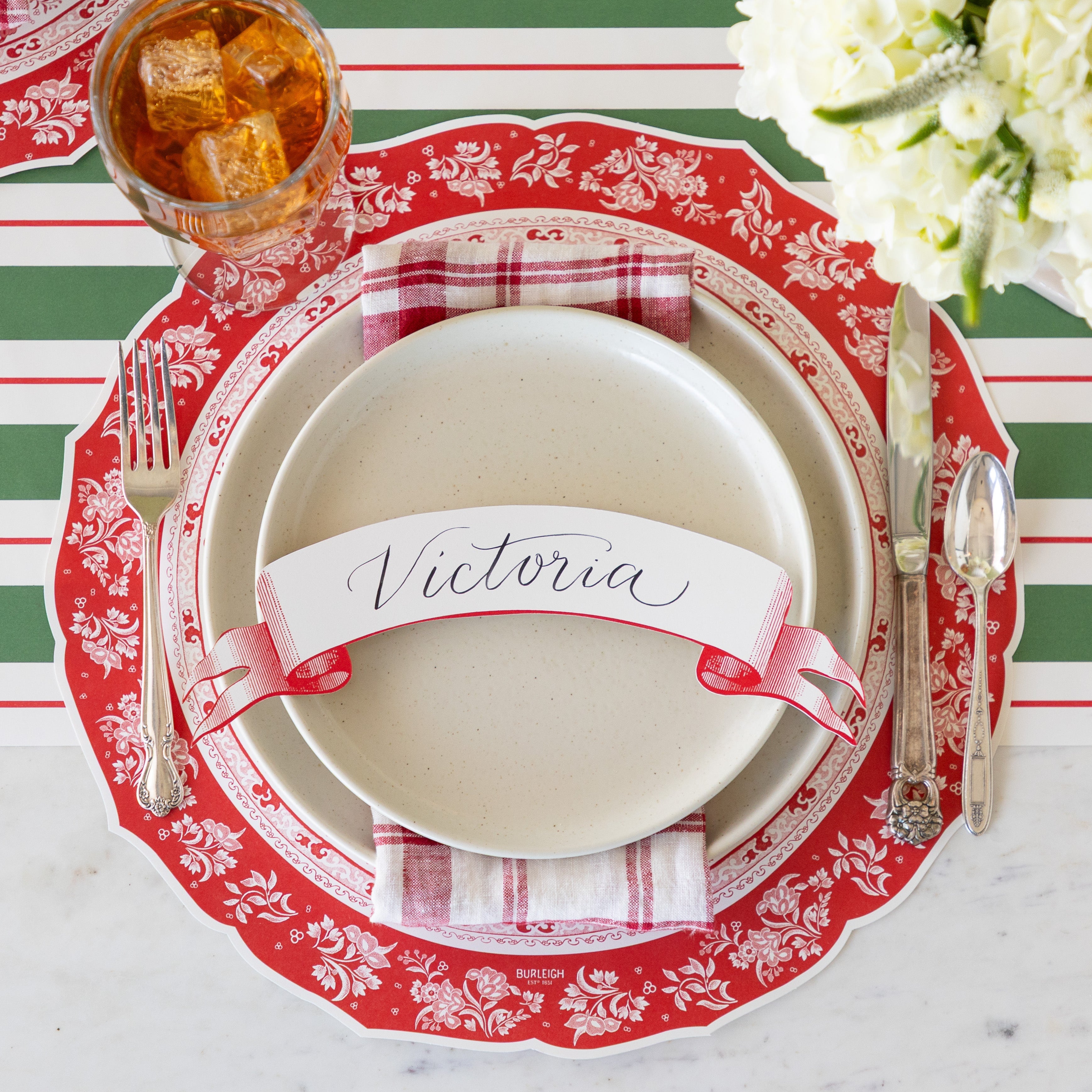 A place setting featuring the Die-cut Red Regal Peacock Placemat, off-white dinner plates with a red-and-white checkered napkin in between and Classic Red Banner Table Accent reading &quot;Victoria&quot; atop, vintage flatware, a glass of tea, and Green &amp; Red Awning Stripe Runner underneath.