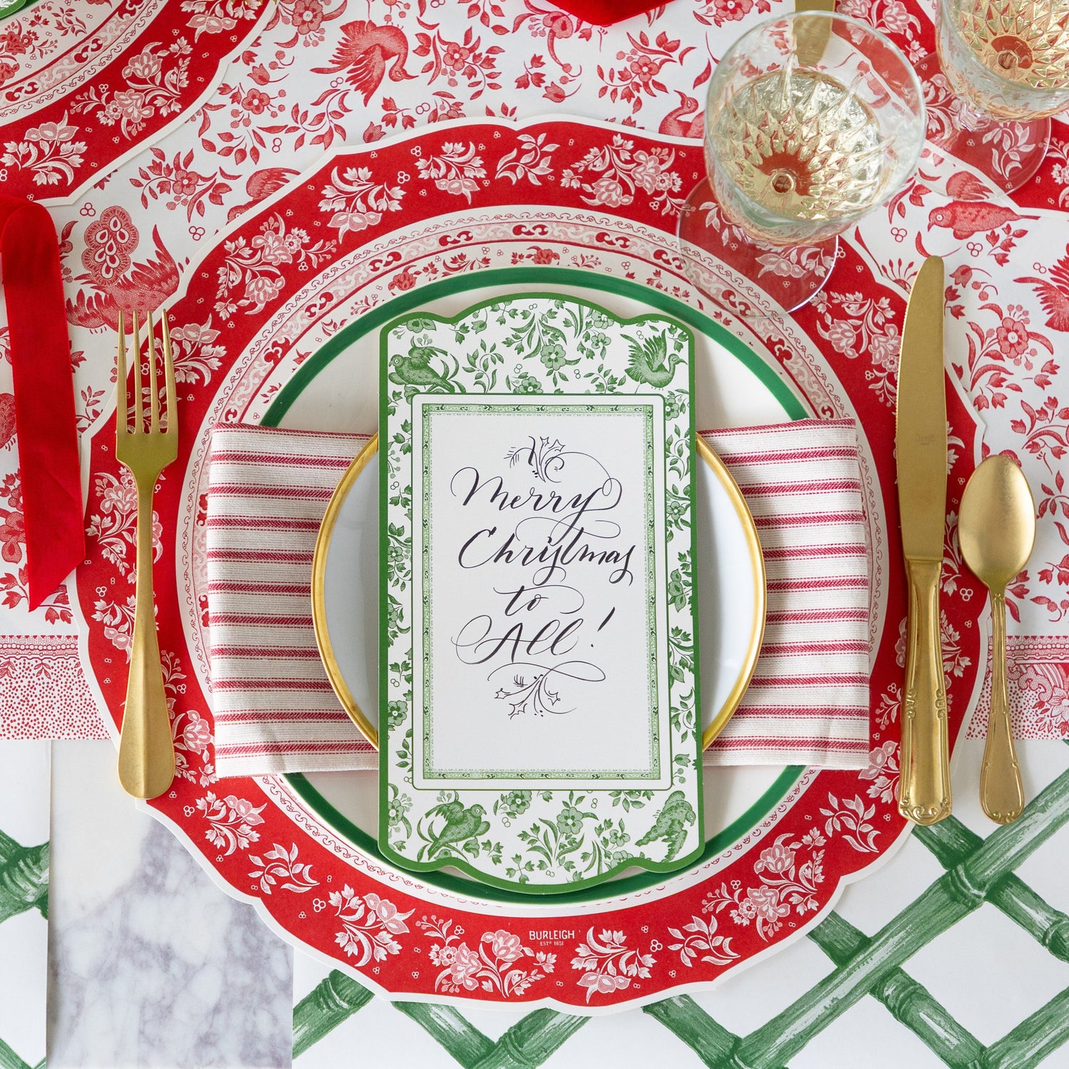 An elegant place setting featuring the Die-cut Red Regal Placemat, green and gold rimmed plates with a red-and-white striped napkin in between, Green Regal Peacock Table Accent reading &quot;Merry Christmas to All!&quot; atop, gold flatware, and Green Lattice Runner and Red Burleigh Peacock Runner underneath.