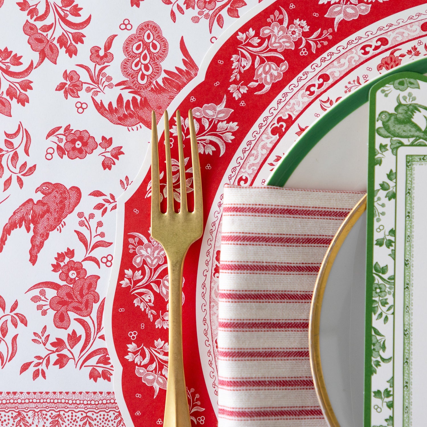 An elegant place setting featuring the Die-cut Red Burleigh Placemat, a gold fork, green-rimmed dinner and gold-rimmed salad plate, red-and-white striped napkin and Red Regal Peacock Runner underneath.